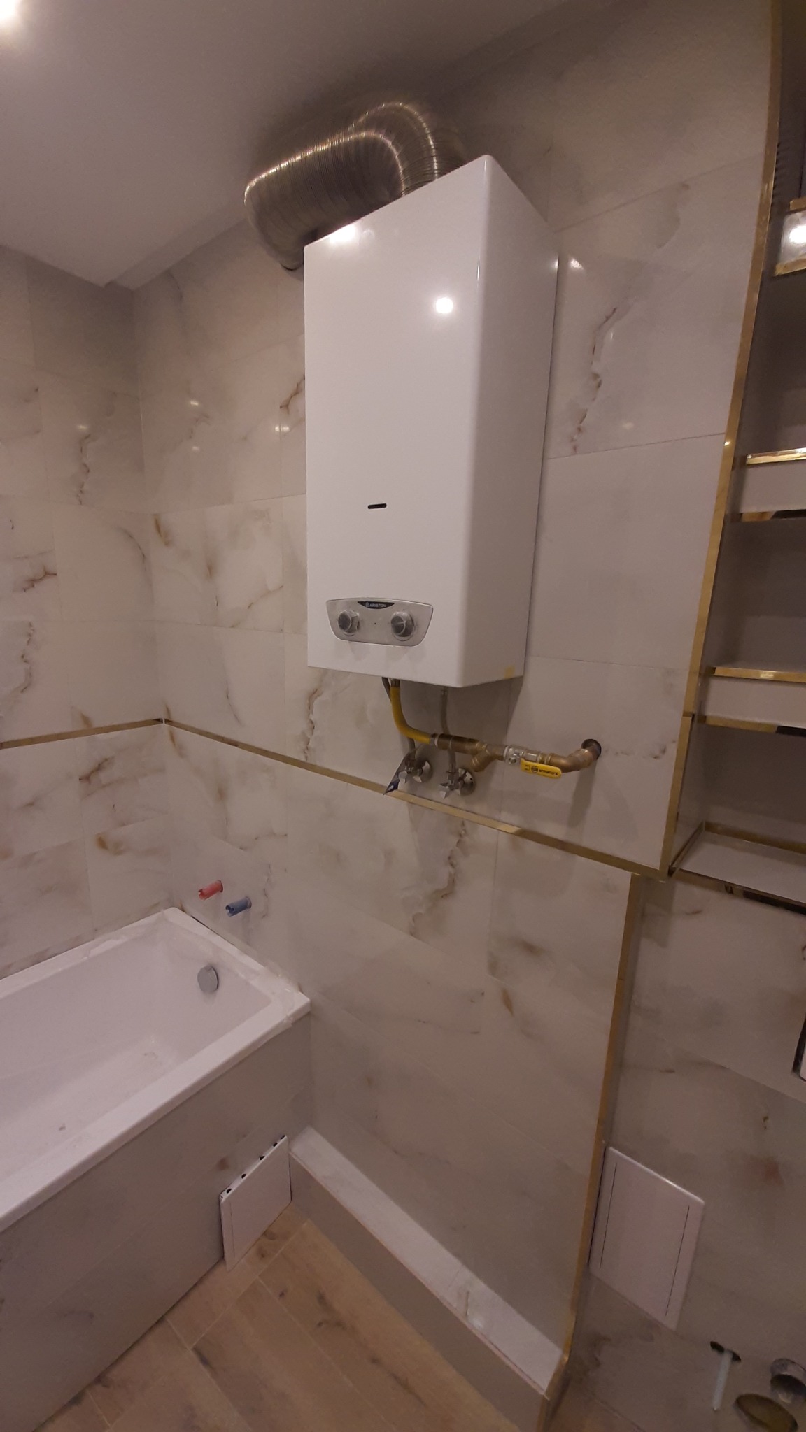 Wykończona łazienka z marmurowymi płytkami, białą wanną i zamontowanym piecykiem gazowym. Widoczne złote akcenty i drewnopodobna podłoga.