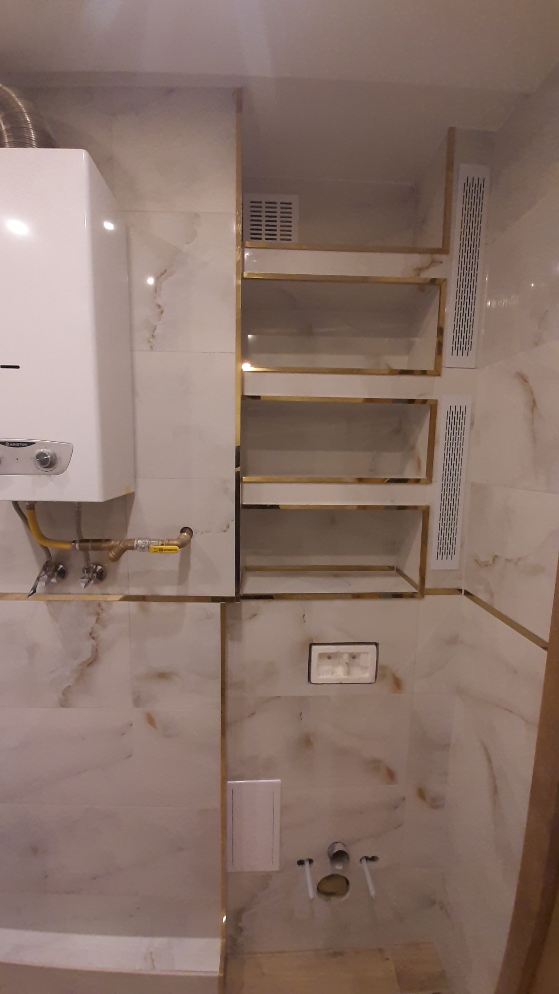 Wykończona łazienka z marmurowymi płytkami, złotymi akcentami i wiszącymi półkami. Widoczny bojler i elementy instalacji wodno-kanalizacyjnej.