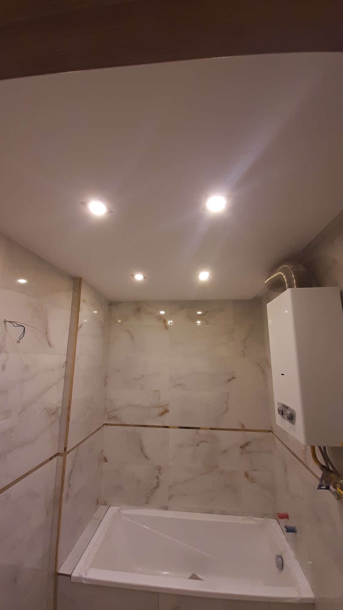 Wykończona łazienka z marmurowymi płytkami, podwieszanym sufitem z oświetleniem punktowym i bojlerem. Widok na wannę i fragment instalacji.