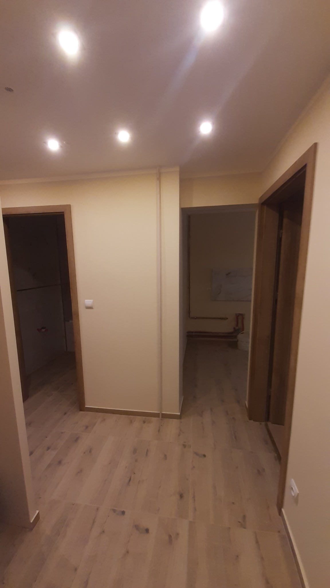 Jasne wnętrze po remoncie: drewnopodobna podłoga, kremowe ściany, dwoje drzwi z drewnianymi ościeżnicami i punktowe oświetlenie sufitowe. Widok na sąsiednie pomieszczenia.