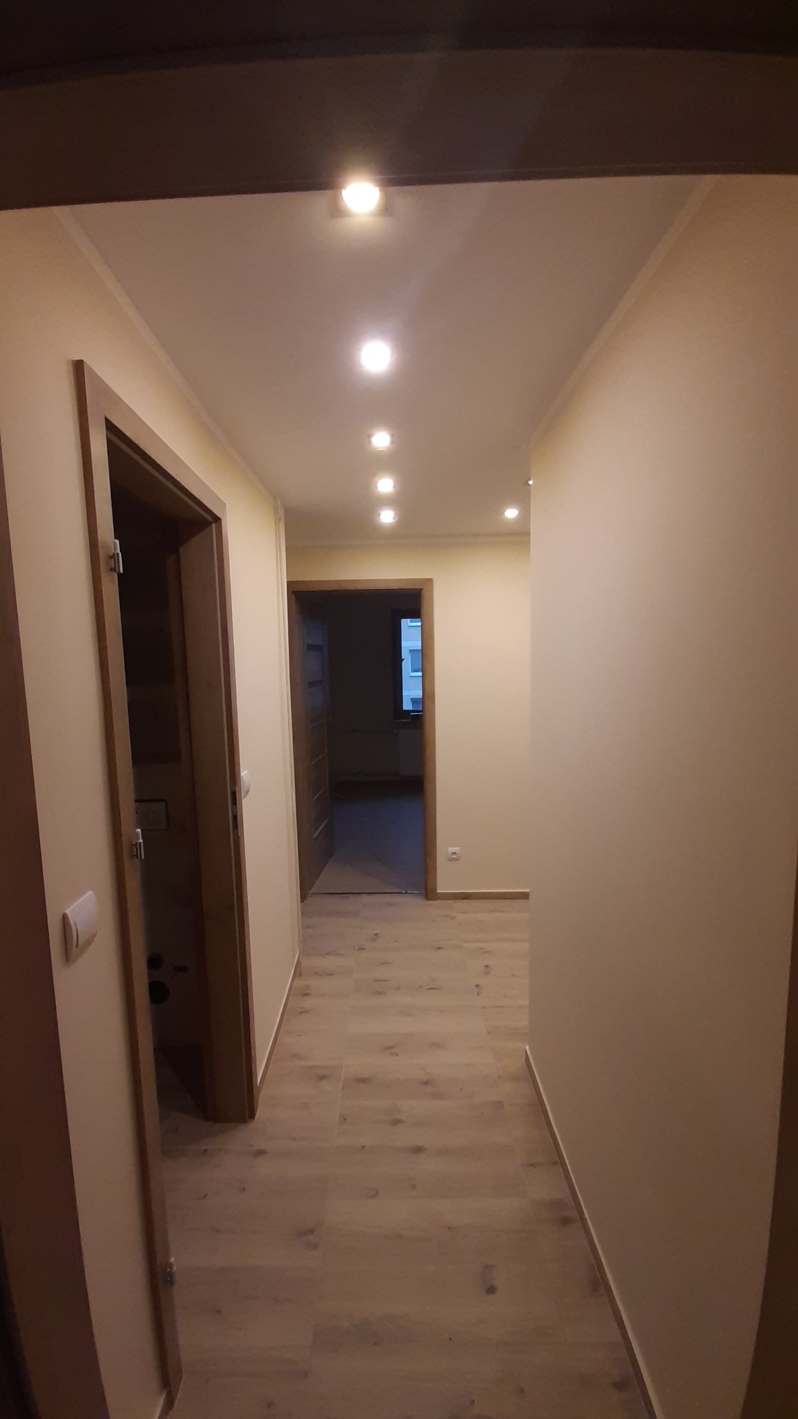 Wykończony korytarz z panelami podłogowymi i oświetleniem punktowym. Widoczne drzwi z drewnianymi ościeżnicami i pomalowane ściany na jasny kolor.