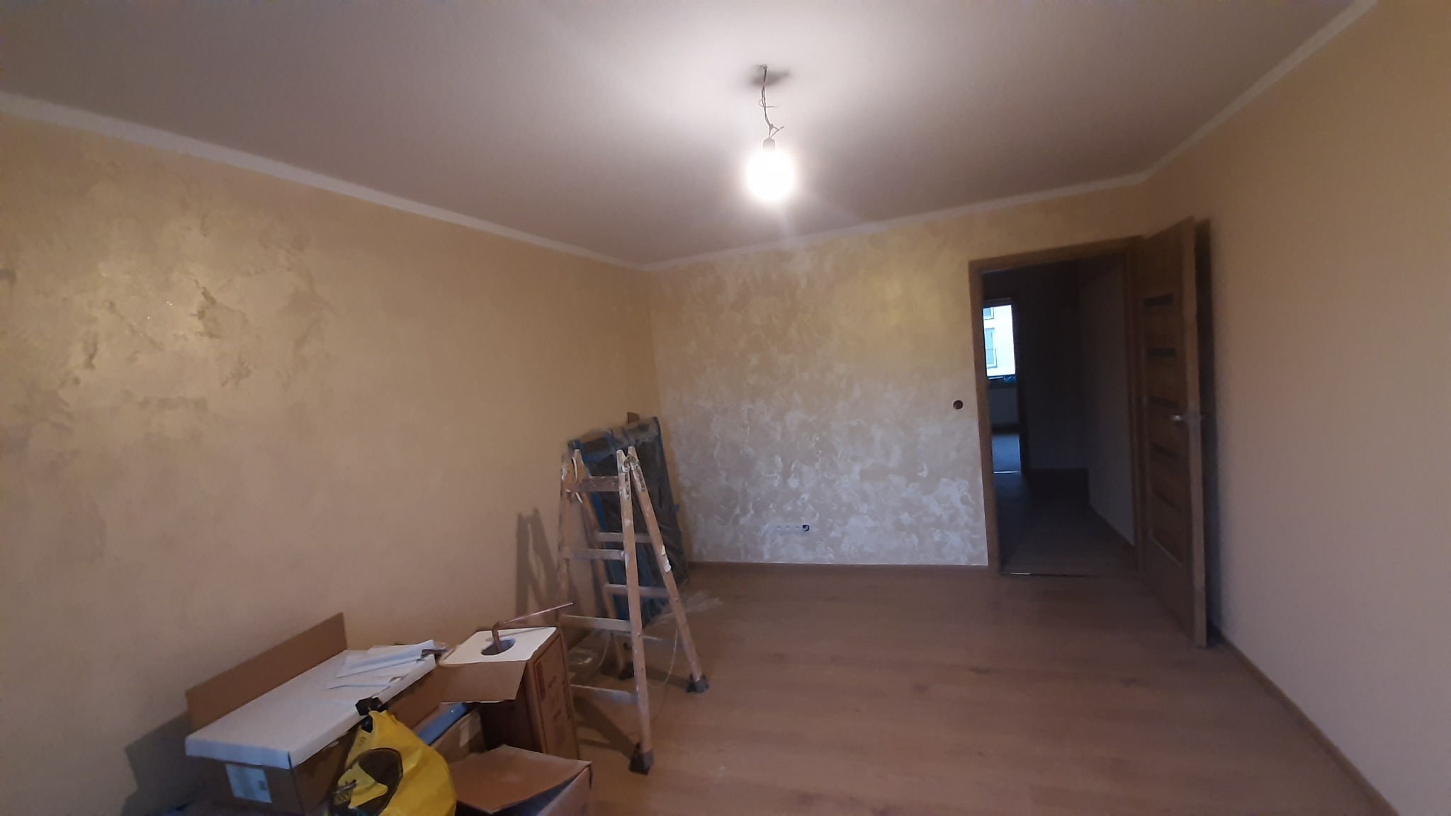 Pokój po malowaniu ścian z efektem dekoracyjnym. Widoczna drabina i akcesoria malarskie. Oświetlenie sufitowe. Otwarta drewniana rama drzwiowa. Podłoga drewniana.