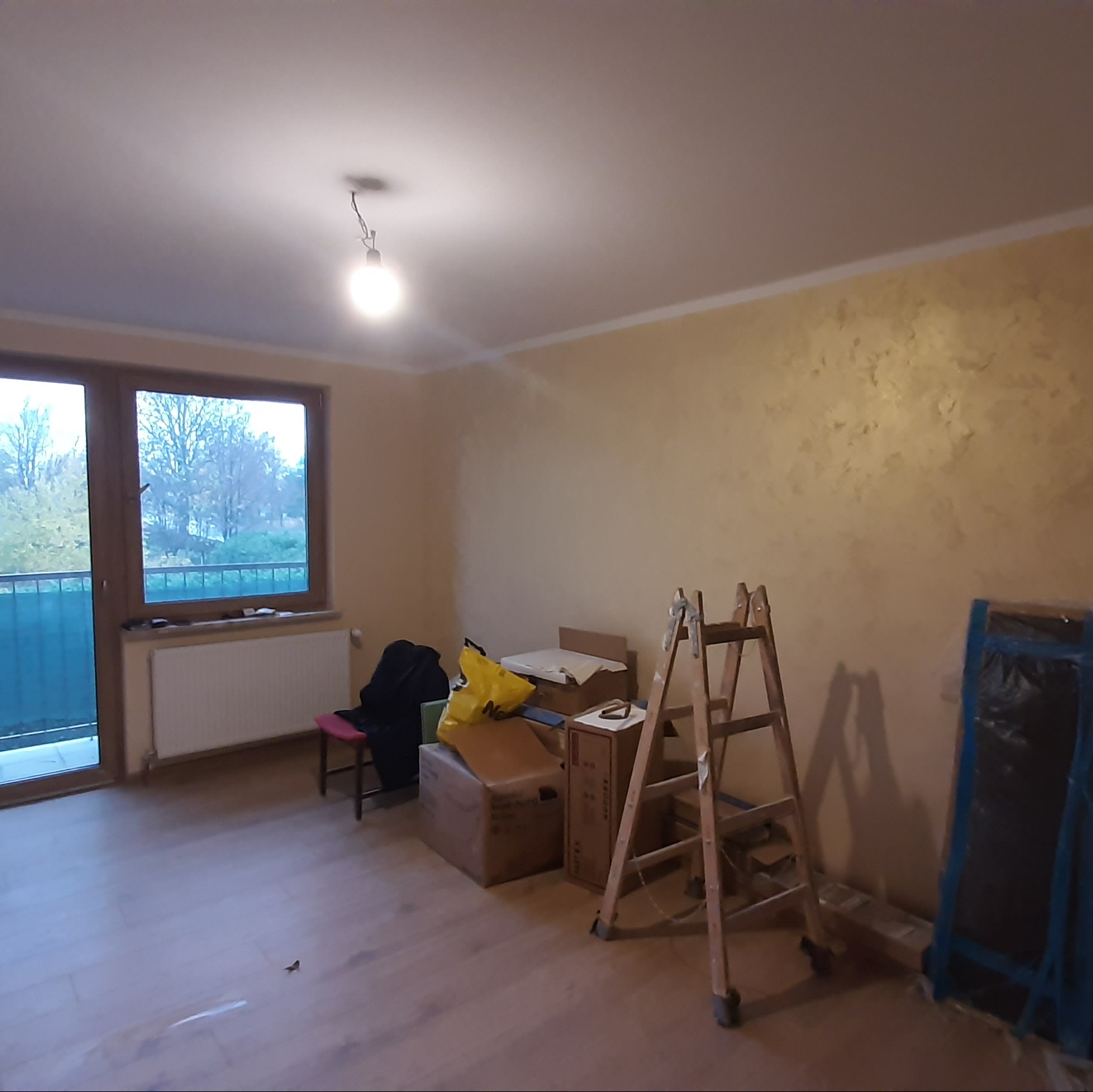 Pokój po malowaniu ścian z efektem dekoracyjnym, widoczne drabiny i zabezpieczone meble. Podłoga wyłożona panelami. Widok na zewnątrz przez okno balkonowe.