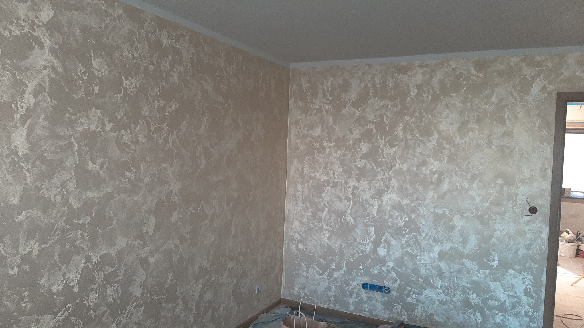 Pokój po dekoracyjnym malowaniu ścian w odcieniach beżu i bieli, z widoczną fakturą. W rogu pomieszczenia widoczne zabezpieczenia podłogi.