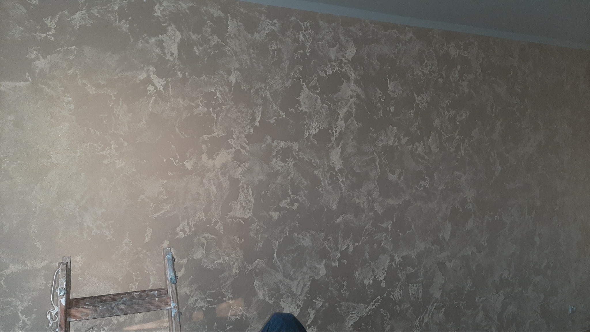 Ściana z dekoracyjną, beżowo-srebrną strukturą, imitującą marmurkowe wzory. W rogu widoczny fragment drabiny malarskiej. Efektowna faktura na gładkiej powierzchni.