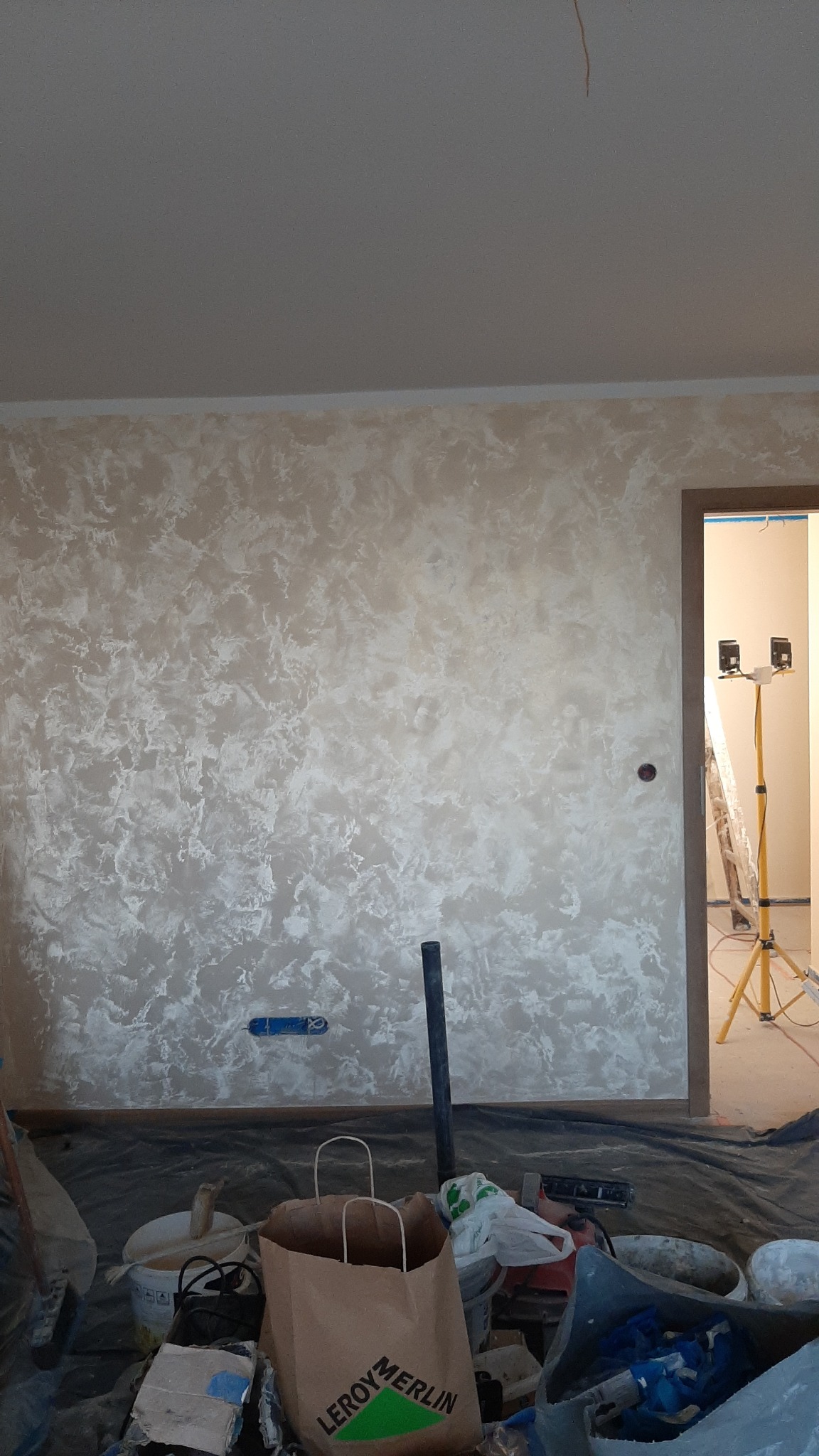 Ściana z dekoracyjną, beżowo-białą strukturą, widoczne narzędzia i akcesoria malarskie, lampa na statywie w przejściu do innego pomieszczenia.
