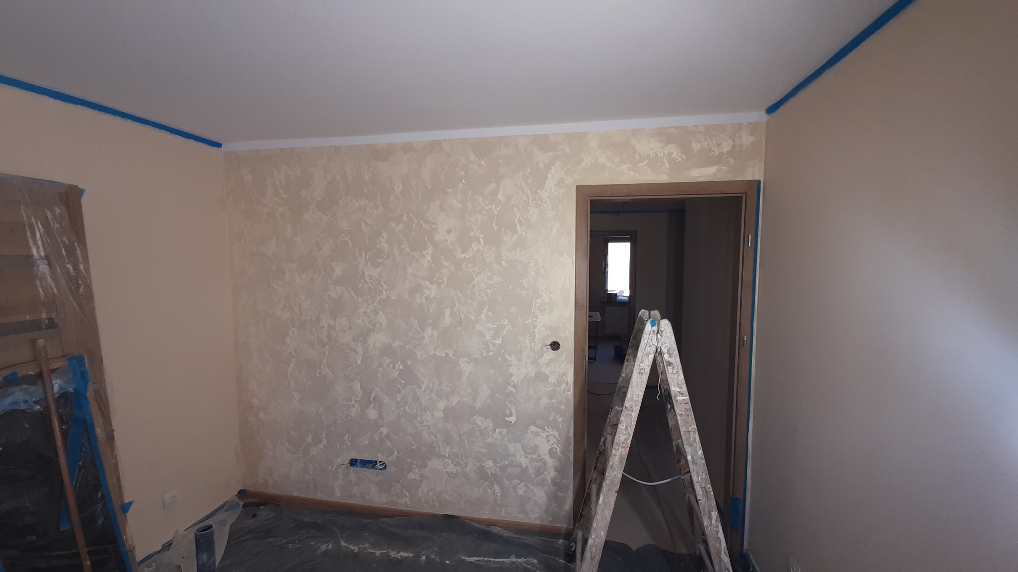 Pokój po dekoracyjnym malowaniu ścian w odcieniach beżu i bieli. Widoczna drabina malarska, zabezpieczona podłoga i taśmy malarskie przy suficie.