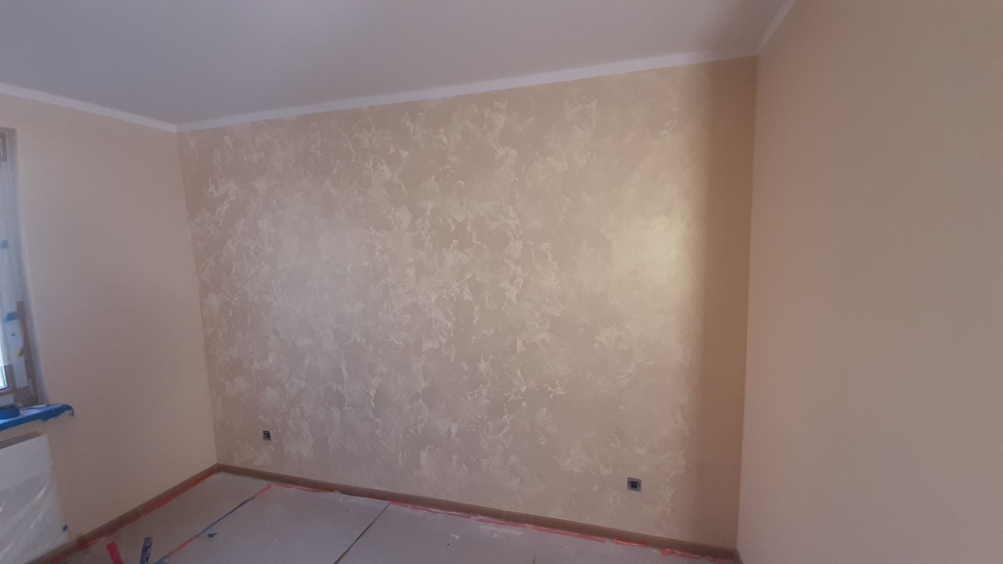 Wykończona ściana z dekoracyjną, beżowo-białą strukturą w pokoju. Widoczne zabezpieczone podłoże i listwa sufitowa. Gniazdko elektryczne na ścianie.