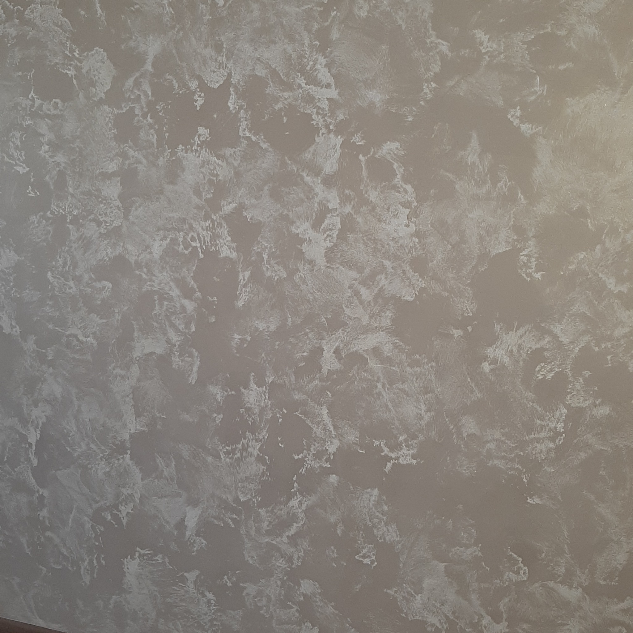 Beżowa ściana z dekoracyjną, nieregularną fakturą w odcieniach bieli i szarości, imitująca efekt marmuru. Widoczne delikatne cieniowanie i subtelne wzory.
