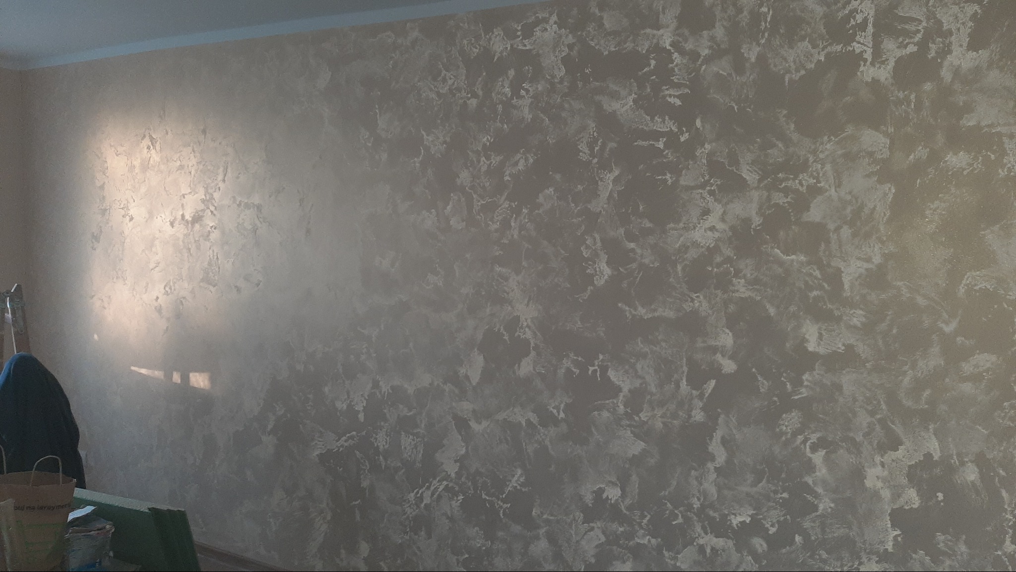 Ściana z dekoracyjną, srebrzystą fakturą, imitującą chmury. Widoczne delikatne refleksy światła. W rogu drabina i przedmioty sugerujące prace wykończeniowe.