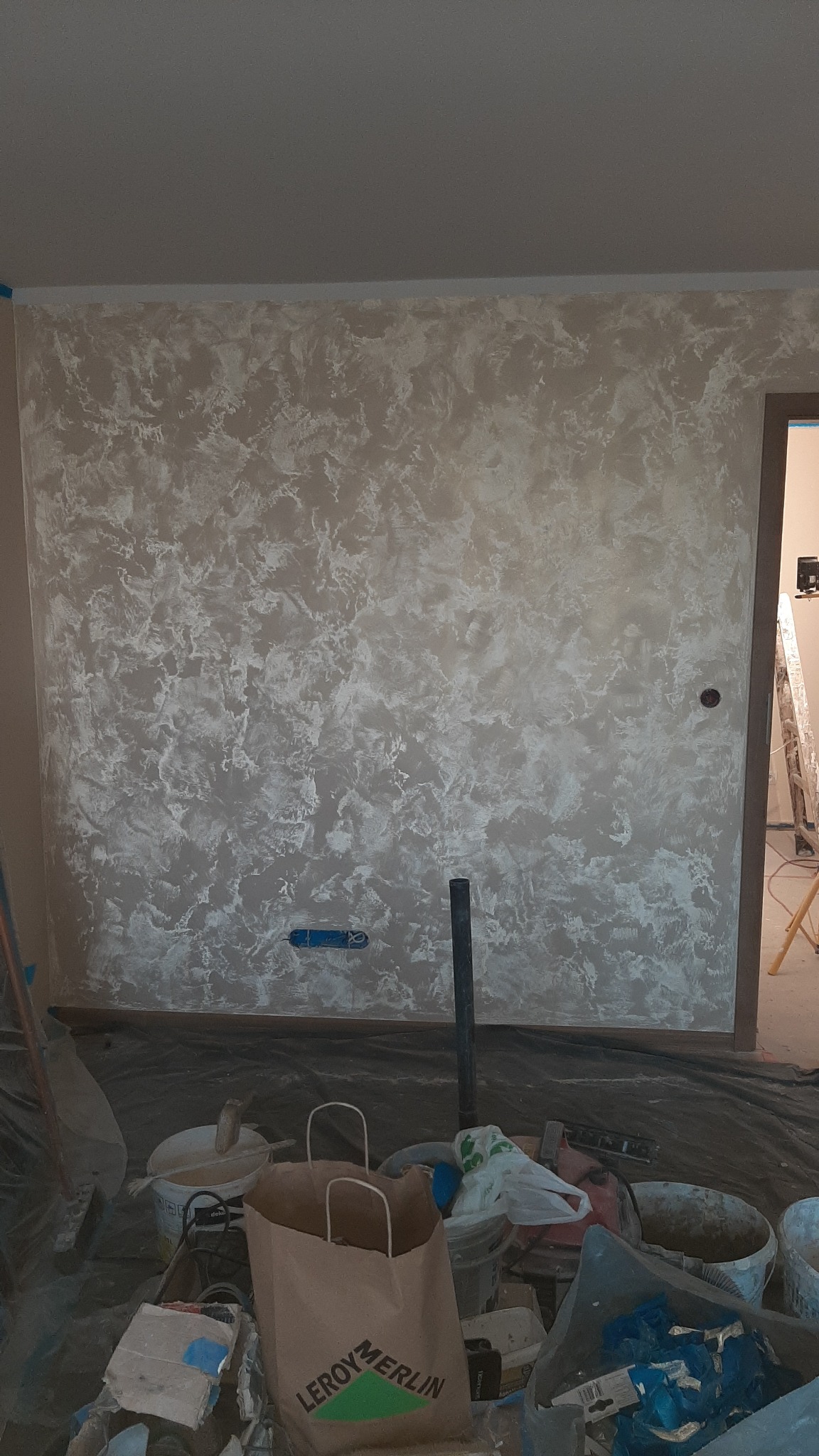 Ściana z dekoracyjną, beżowo-białą strukturą, widoczne narzędzia malarskie i torba Leroy Merlin, sugerujące prace wykończeniowe wnętrz w trakcie realizacji.