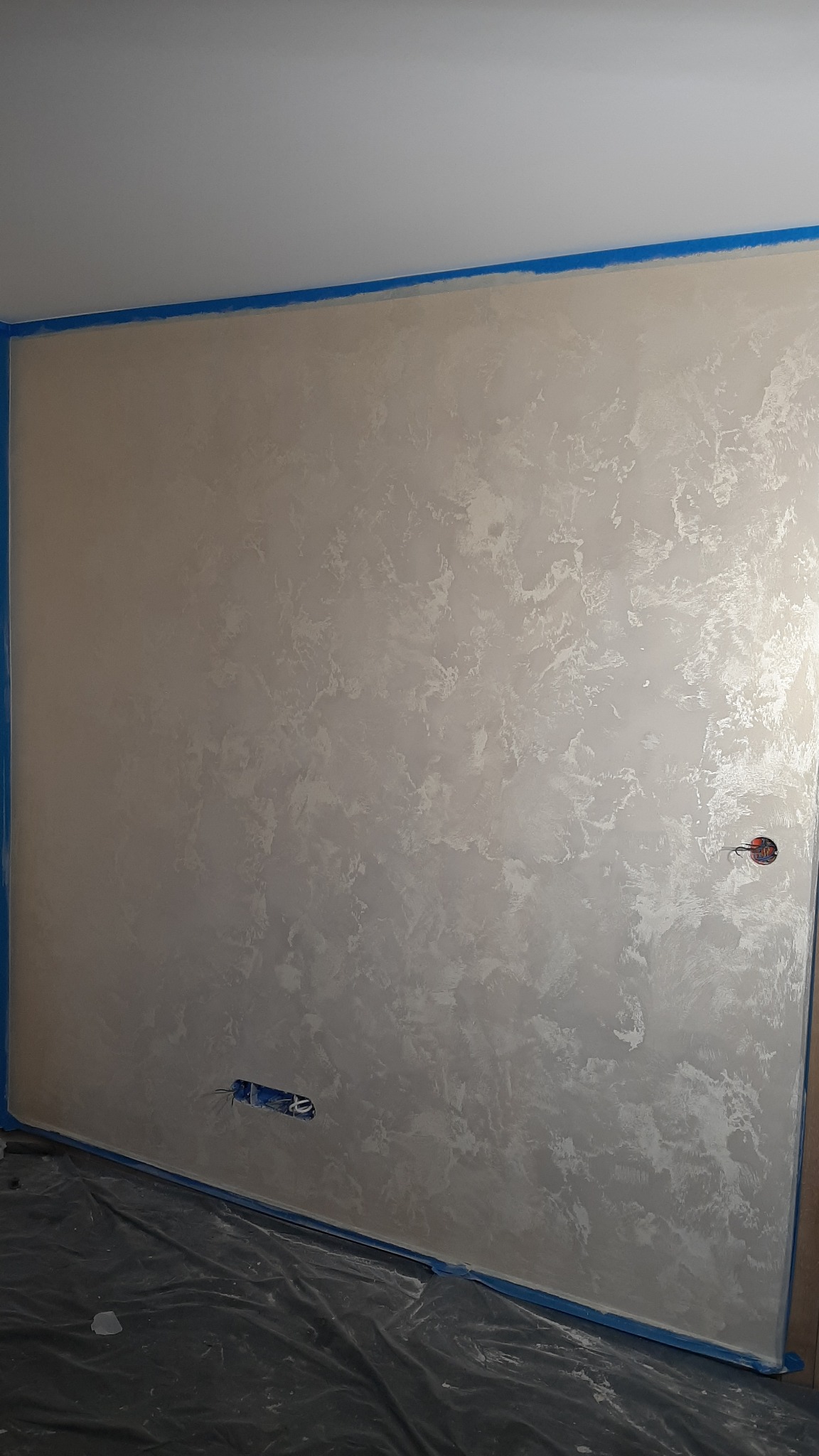 Ściana z dekoracyjną, srebrzystą fakturą, zabezpieczona niebieską taśmą malarską przy suficie i podłodze. Widoczne gniazdka elektryczne. Podłoga zabezpieczona folią.