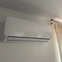 Biała klimatyzacja marki AUX zamontowana na ścianie, widoczne ż&oacute;łte zasłony w tle, oświetlenie dzienne.