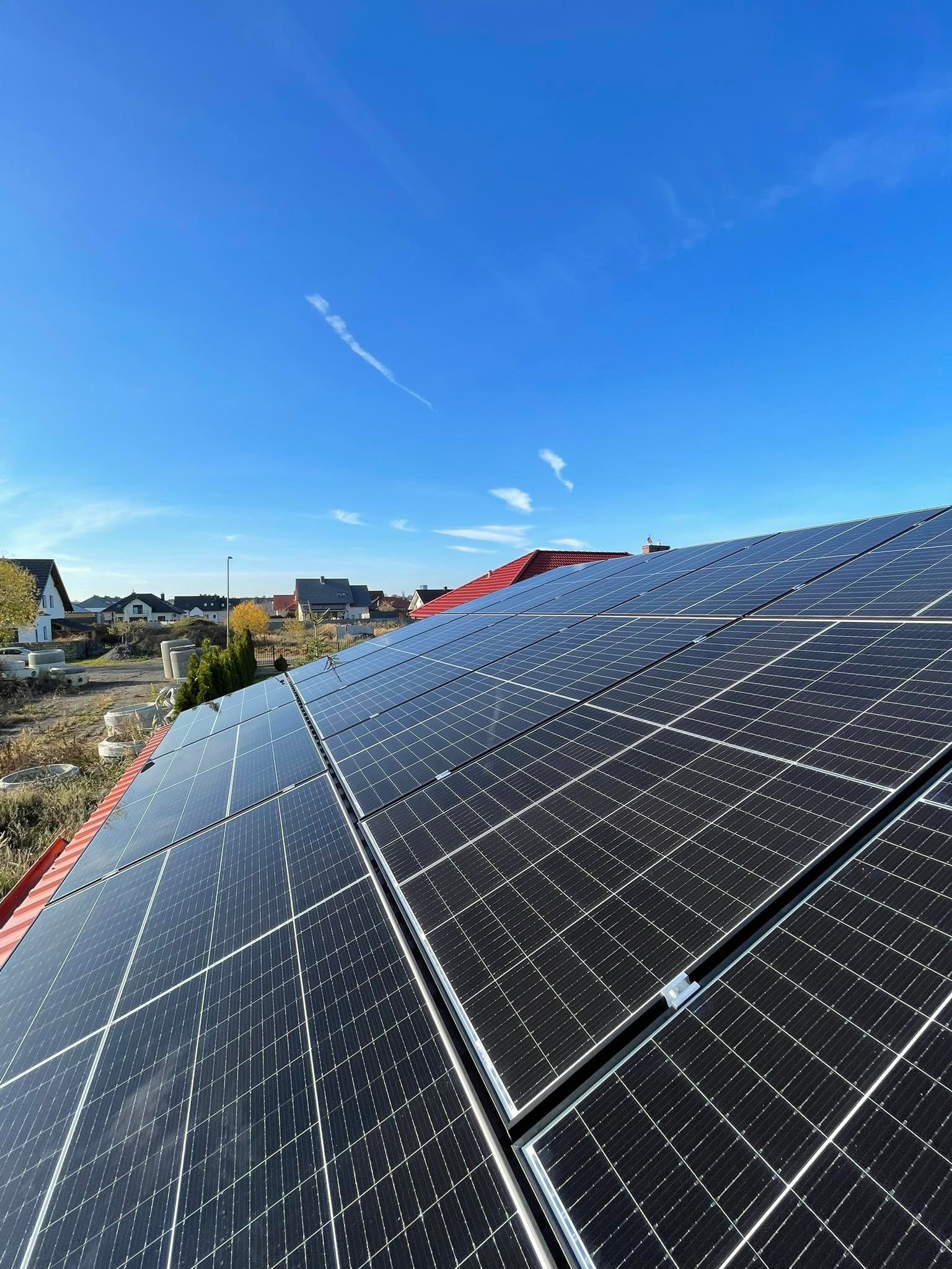 Panele fotowoltaiczne na czerwonym dachu domu jednorodzinnego w słoneczny dzień, widoczne białe smugi na błękitnym niebie