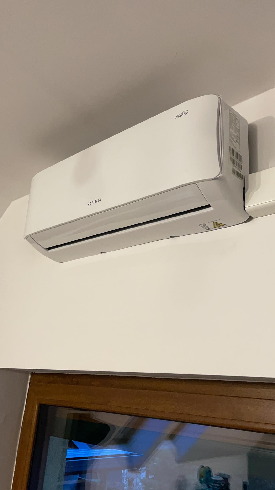 Biała jednostka wewnętrzna klimatyzacji Rotenso zamontowana na ścianie, widok z dołu, z fragmentem ramy okiennej w tle.