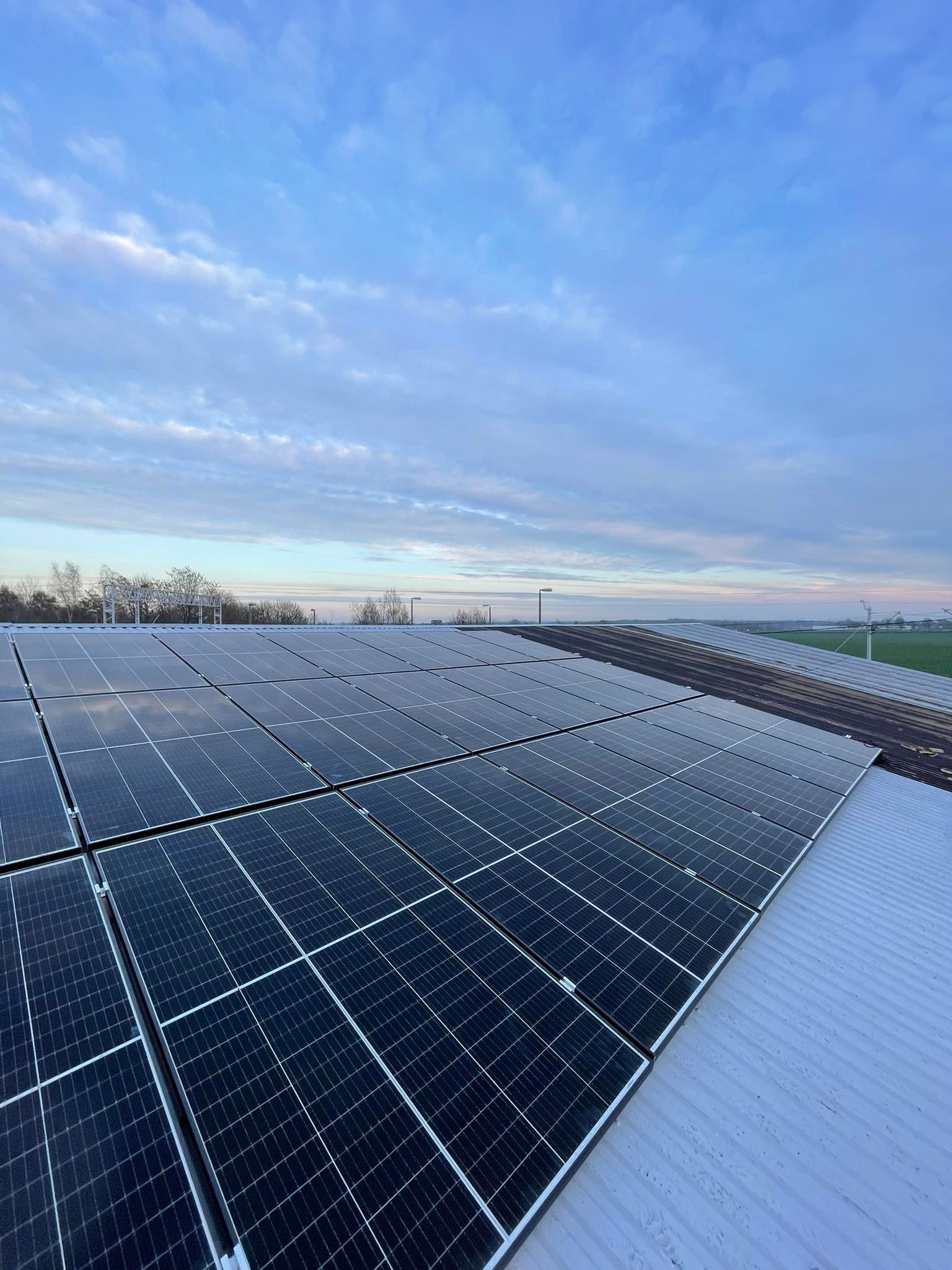 Rząd paneli słonecznych na dachu budynku, widok z góry pod lekkim kątem, z widocznym horyzontem i niebem z chmurami.