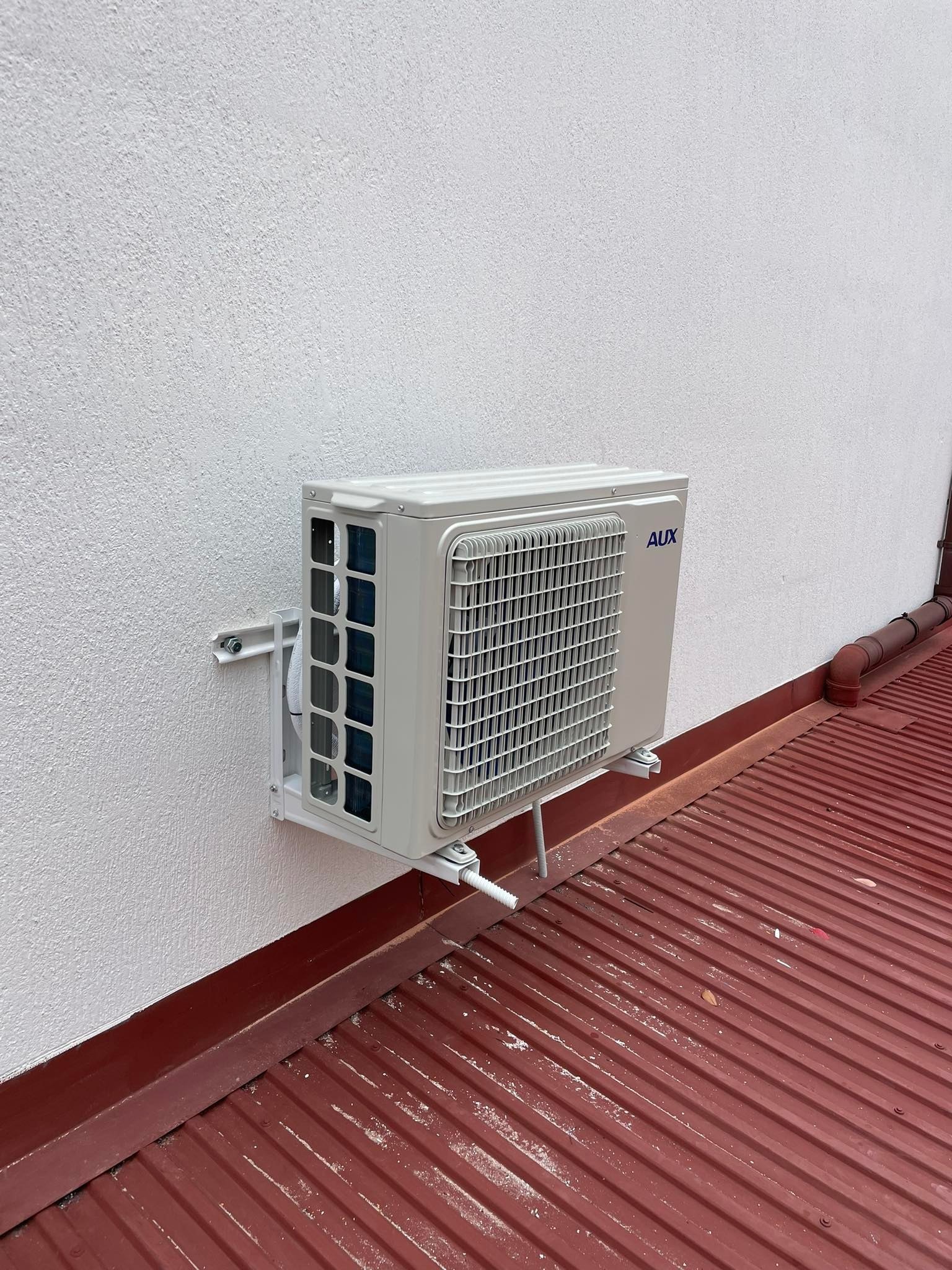 Zewnętrzna jednostka klimatyzacji marki AUX zamontowana na białej, chropowatej ścianie budynku, z widoczną instalacją odprowadzającą skropliny, na tle pomarańczowej blachy dachowej.
