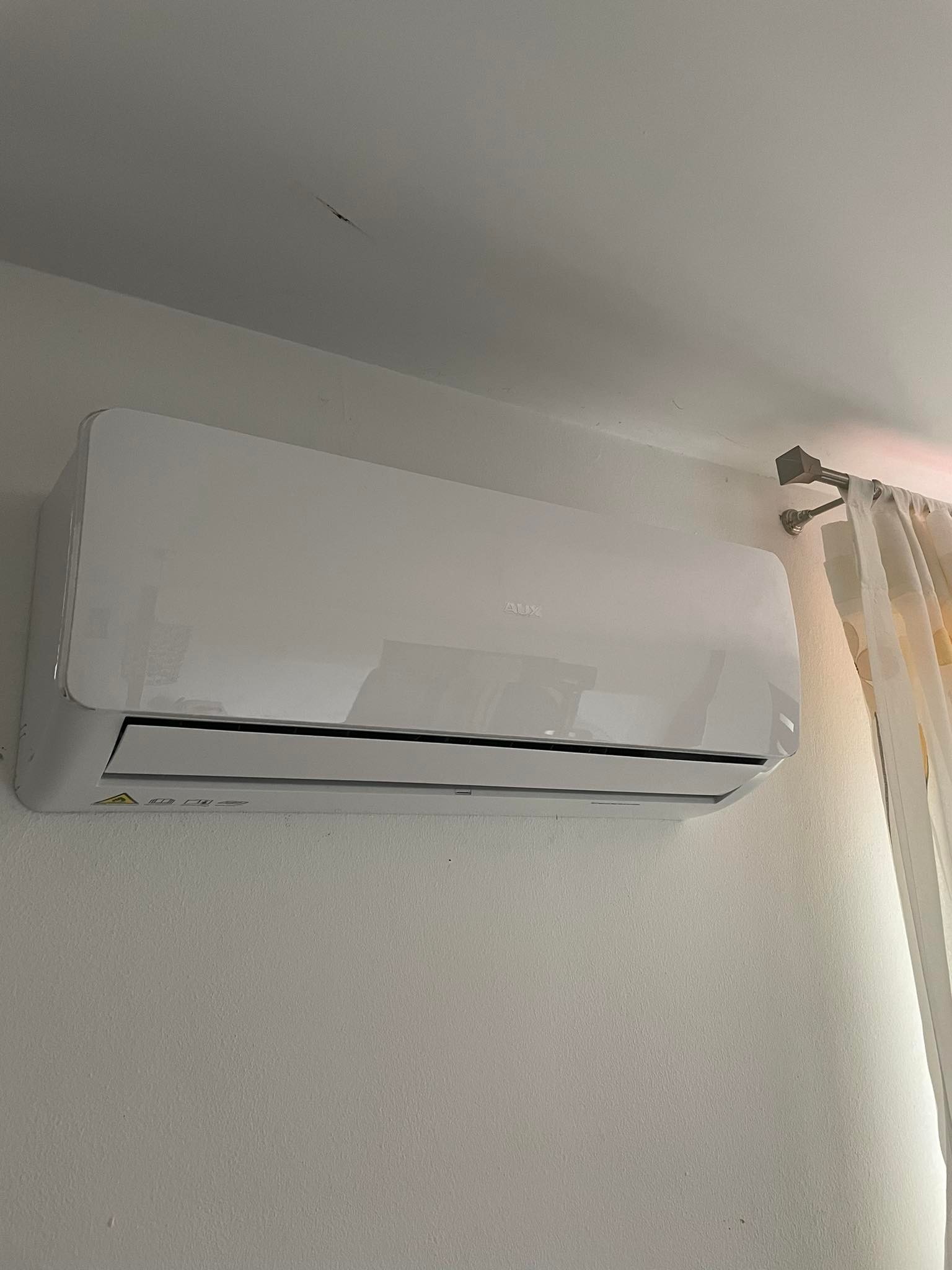 Biała klimatyzacja marki AUX zamontowana na ścianie, widoczne żółte zasłony w tle, oświetlenie dzienne.
