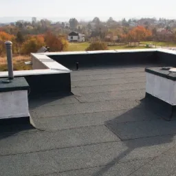 Wykończony płaski dach pokryty papą termozgrzewalną z kominkami wentylacyjnymi i attyką, widok na miasto w tle.