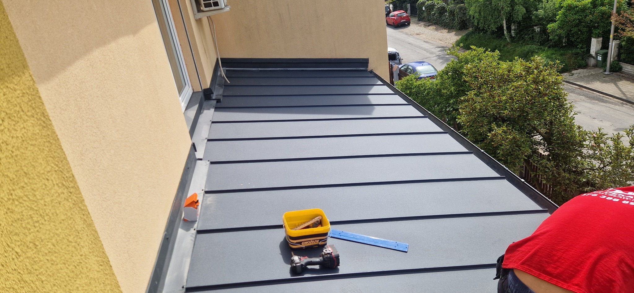 Nowy, grafitowy dach z blachy na rąbek stojący, widok z góry na narzędzia: wiertarko-wkrętarka, pojemnik na gwoździe i miarka; w tle zabudowa miejska z zielenią.