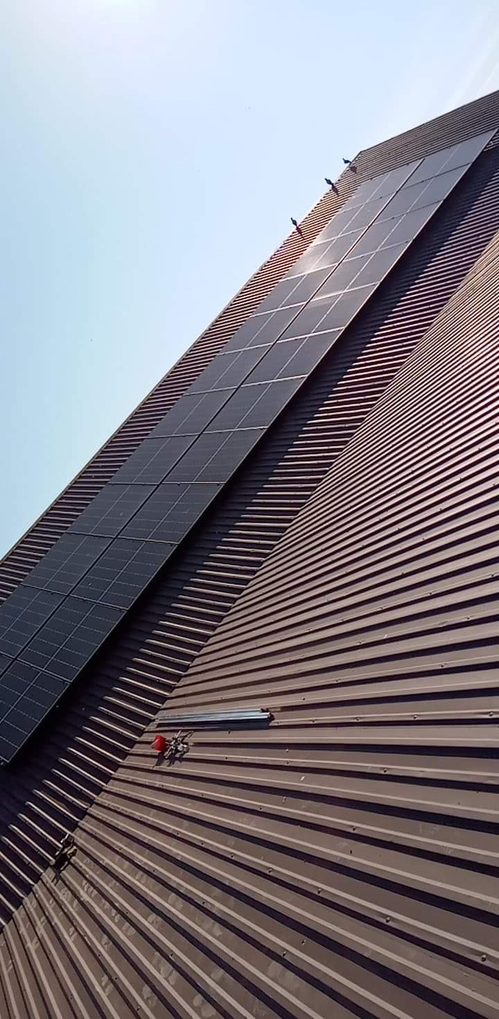 Panele fotowoltaiczne zamontowane na brązowym, metalowym dachu z widocznymi elementami montażowymi i trzema ptakami siedzącymi na górnej krawędzi paneli, z jasnym niebem w tle.