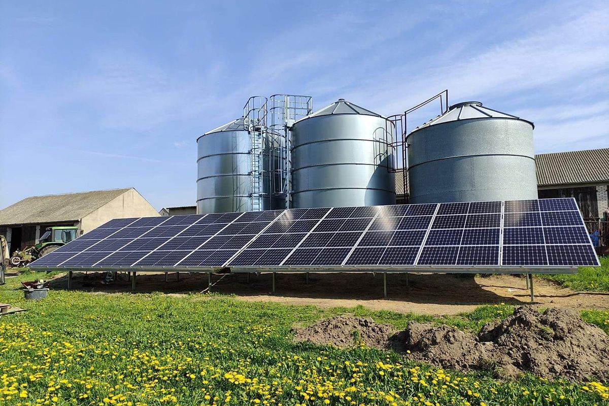 Panel fotowoltaiczny zamontowany na łące z widokiem na silosy zbożowe i budynki gospodarcze w tle.