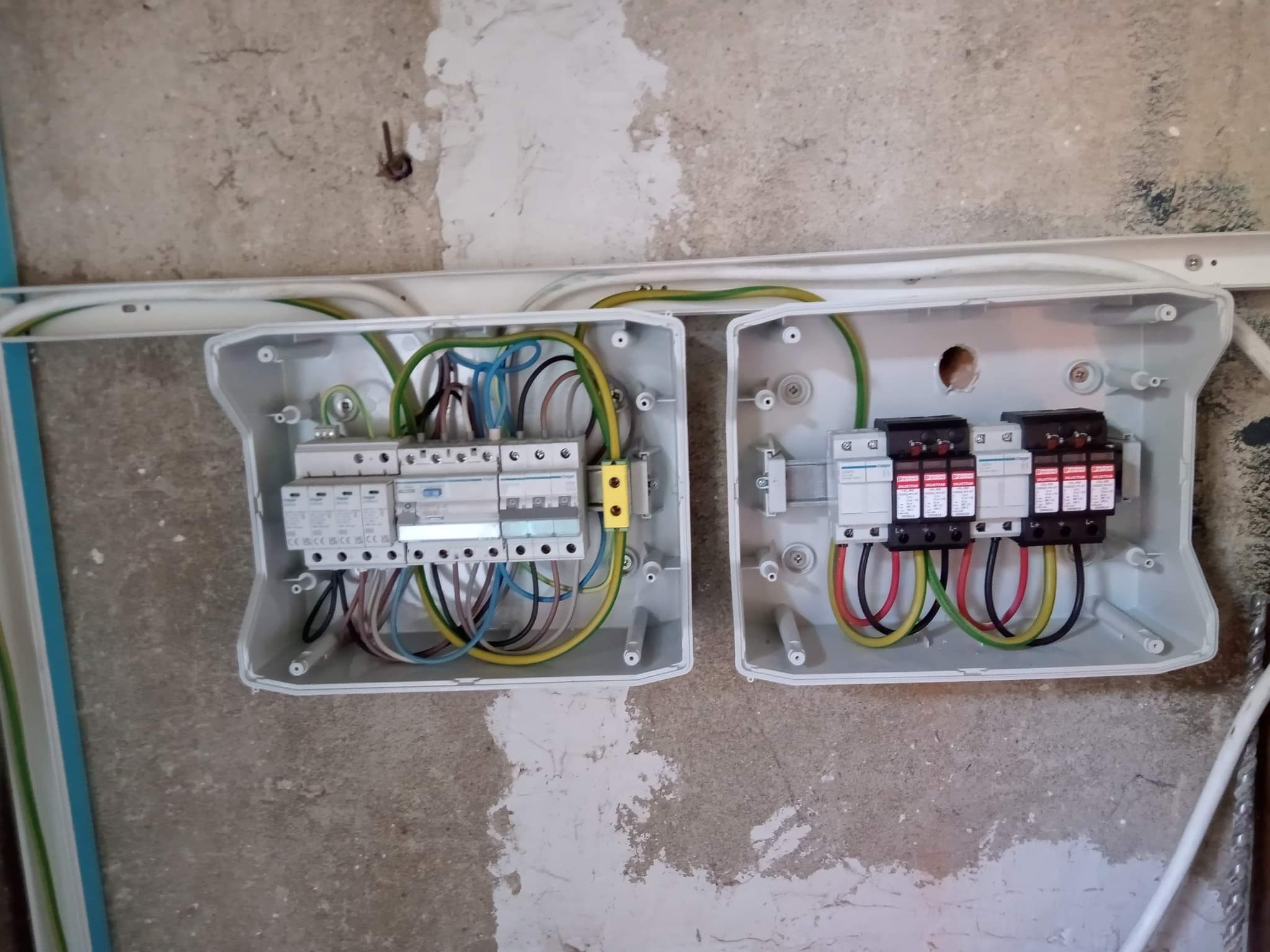 Dwa otwarte panele elektryczne z widocznymi przewodami i bezpiecznikami zamontowane na surowej ścianie podczas instalacji.