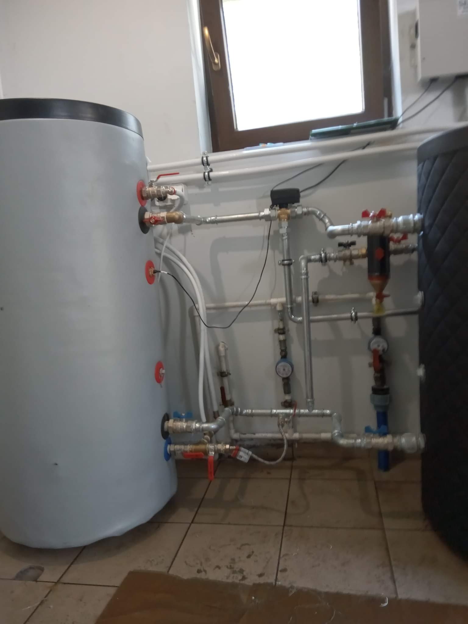 Dwa akumulatory ciepła, jeden szary i jeden czarny, połączone rurami i zaworami w pomieszczeniu z oknem, widoczne manometry i filtr.
