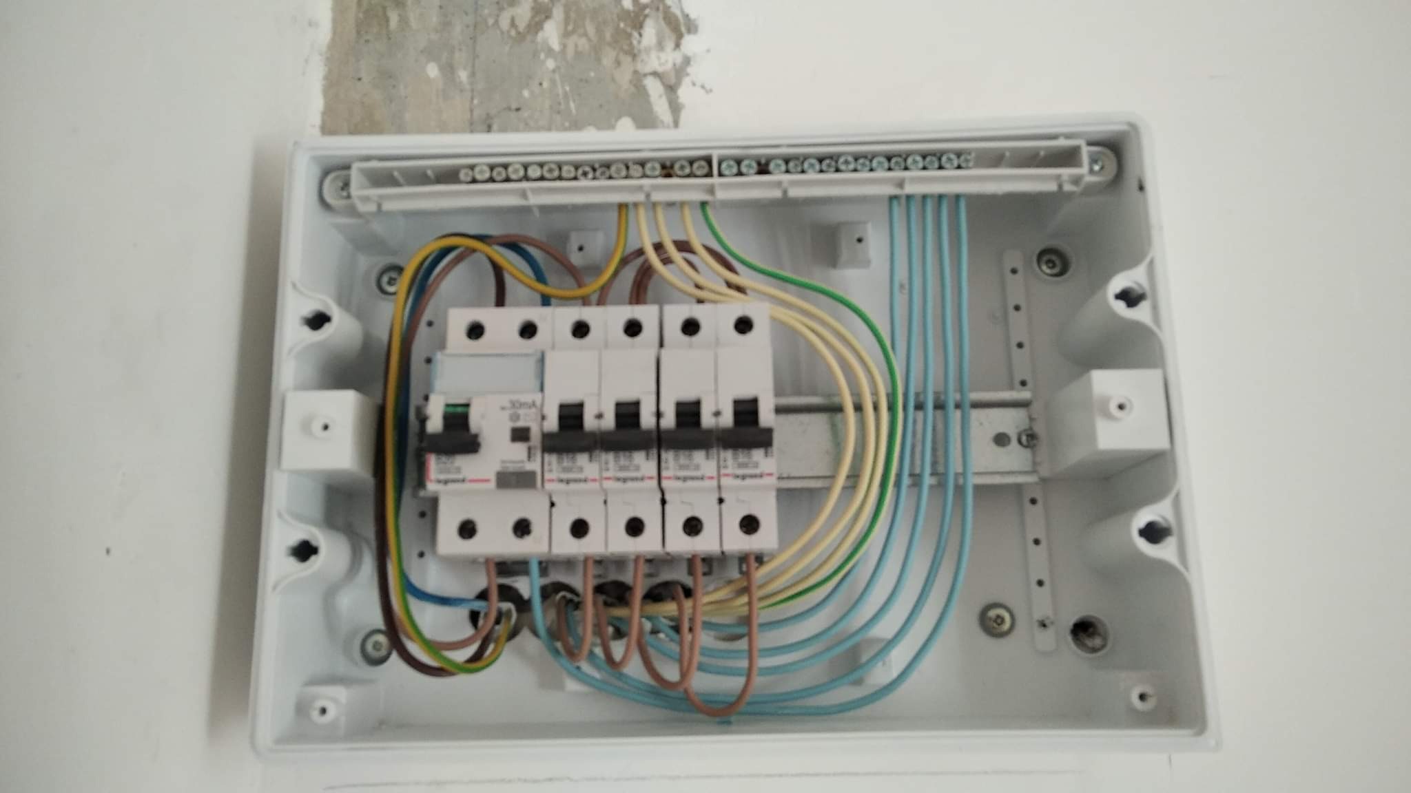 Otwarta biała skrzynka elektryczna z zamontowanymi bezpiecznikami i podłączonymi przewodami w różnych kolorach, przymocowana do ściany z widocznymi śladami po naprawach.