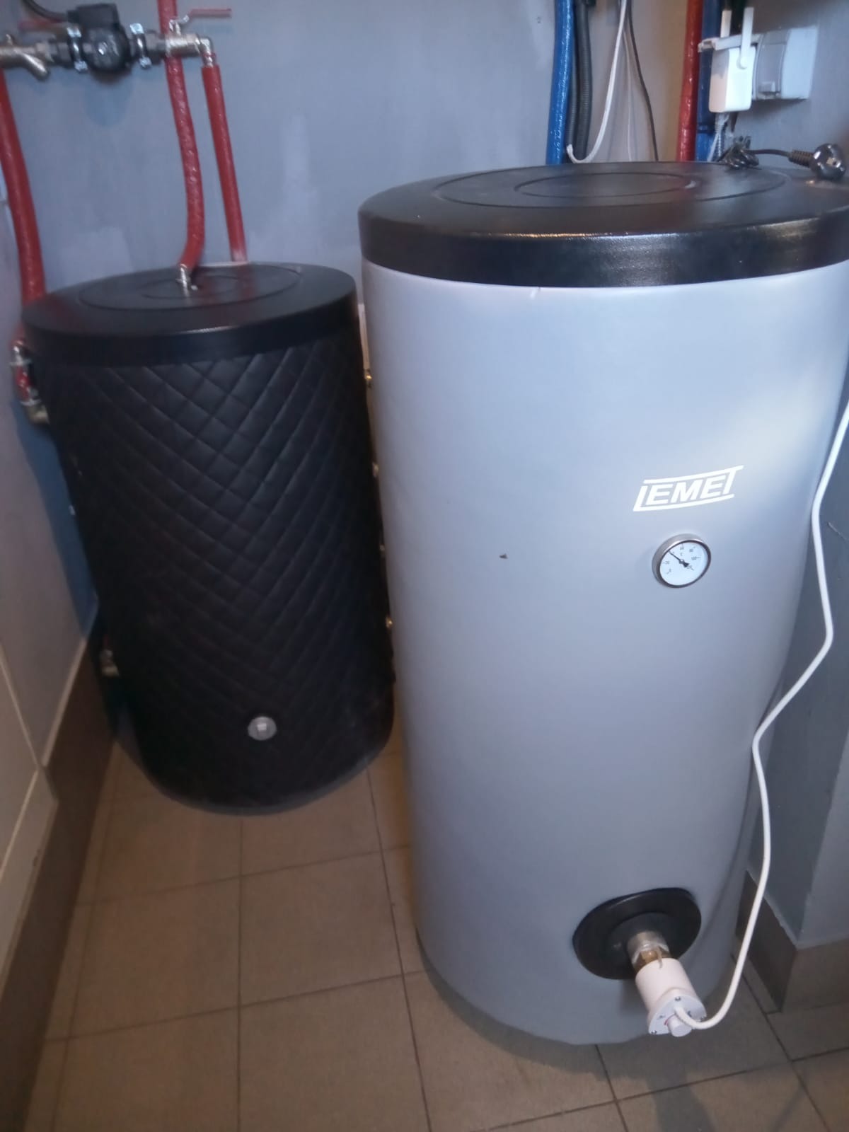 Dwa cylindryczne zbiorniki w pomieszczeniu: czarny z pikowaną teksturą i szary z manometrem, oba podłączone do instalacji.