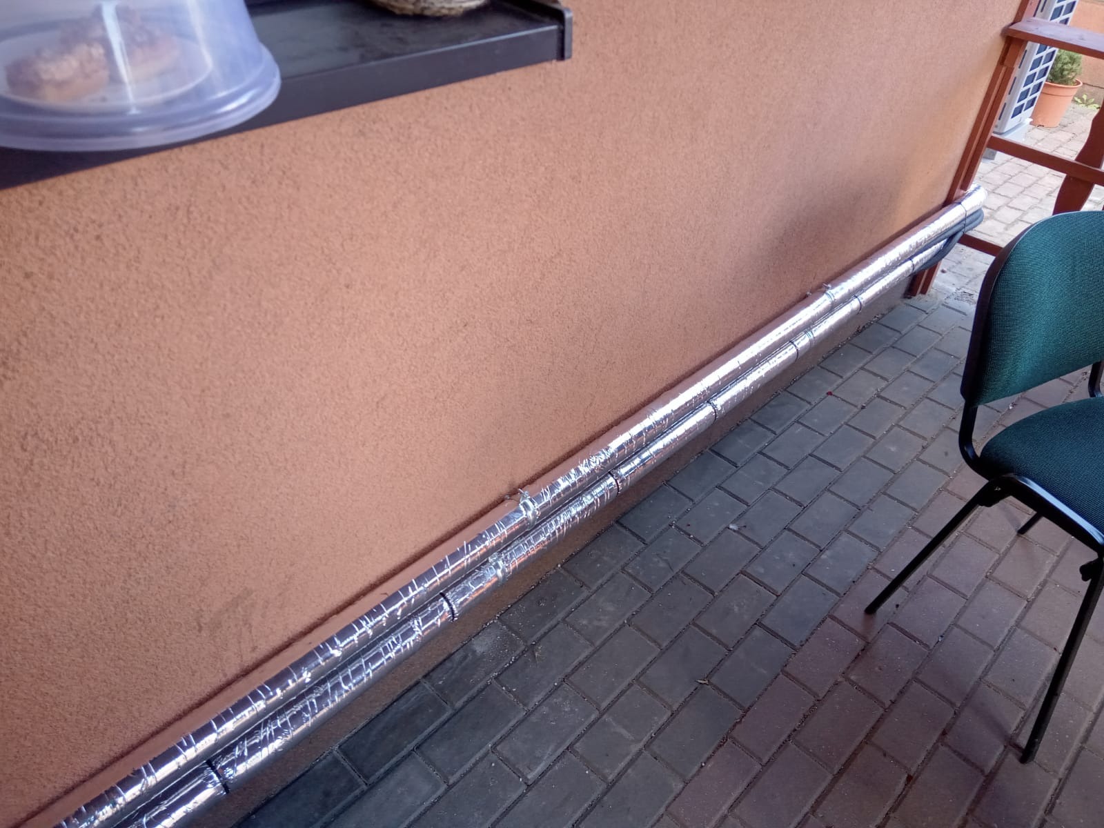 Dwie równoległe, srebrne rury izolacyjne zabezpieczone taśmą, biegnące wzdłuż ściany budynku z beżowym tynkiem, na tle szarej kostki brukowej i fragmentu krzesła.
