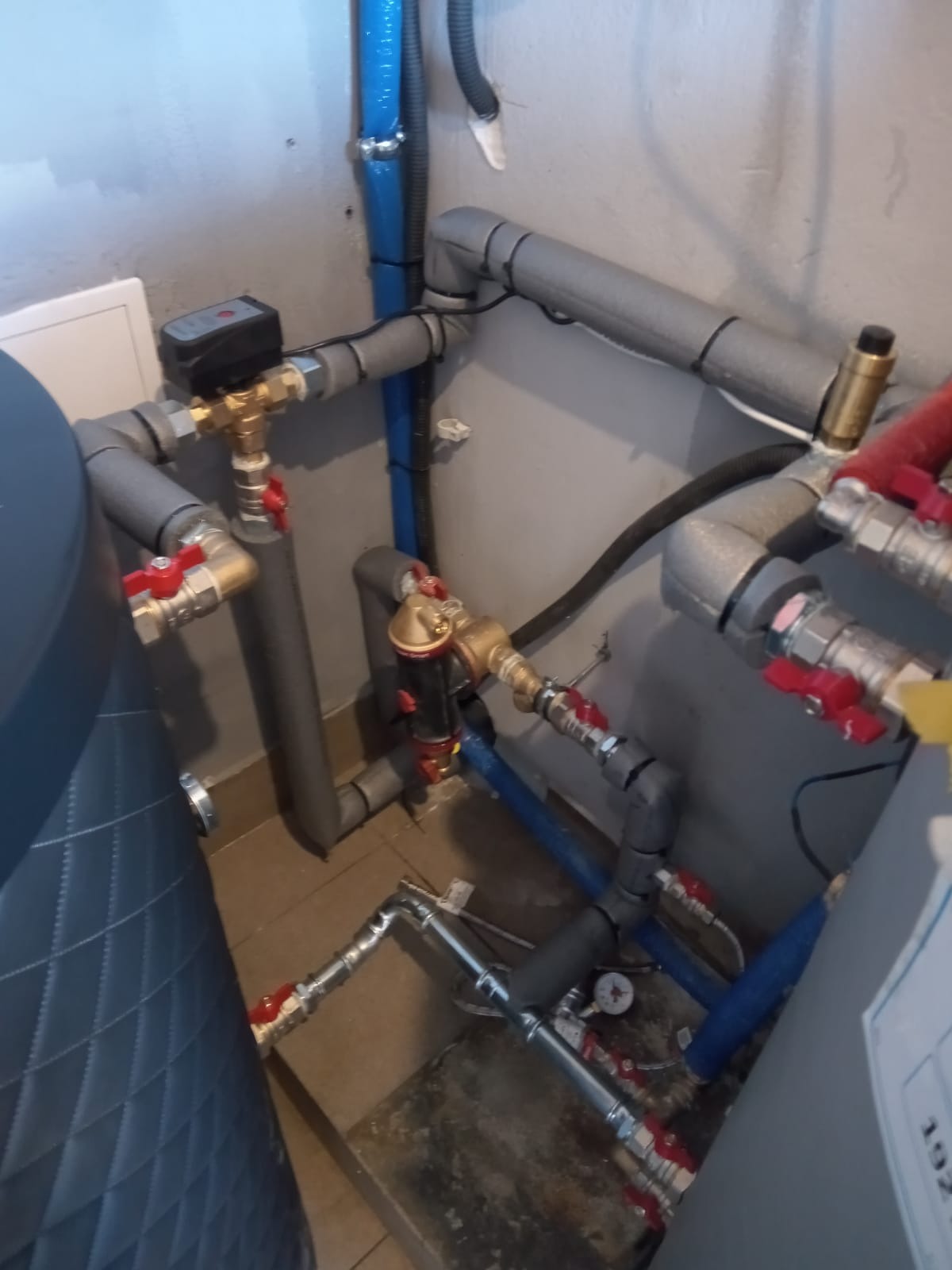 Złożony system rur z zaworami, manometrem i elementami izolacji termicznej w szarej kotłowni, widok z góry na instalację wodną.