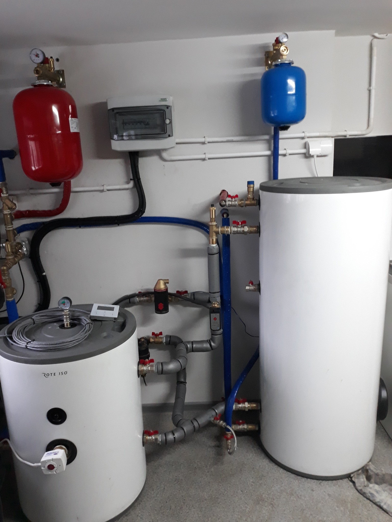 Dwa białe zasobniki ciepłej wody użytkowej z podłączonymi rurami i zaworami, czerwony i niebieski zbiornik wyrównawczy, szara skrzynka elektryczna na ścianie.