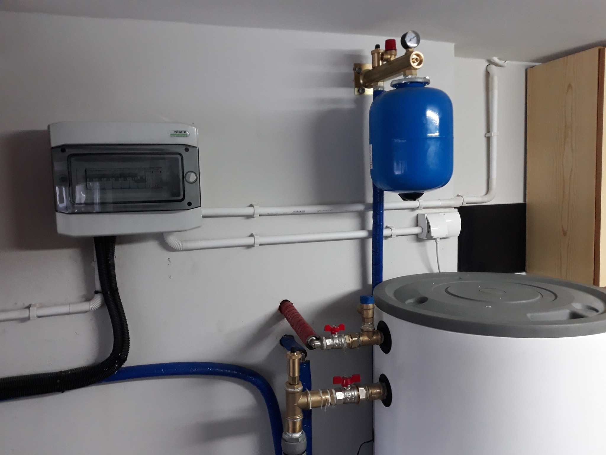 Zamknięty obieg wody grzewczej: biały zasobnik, niebieskie naczynie wzbiorcze, rury miedziane z zaworami, manometr, zabezpieczenia i rozdzielnia elektryczna na ścianie.