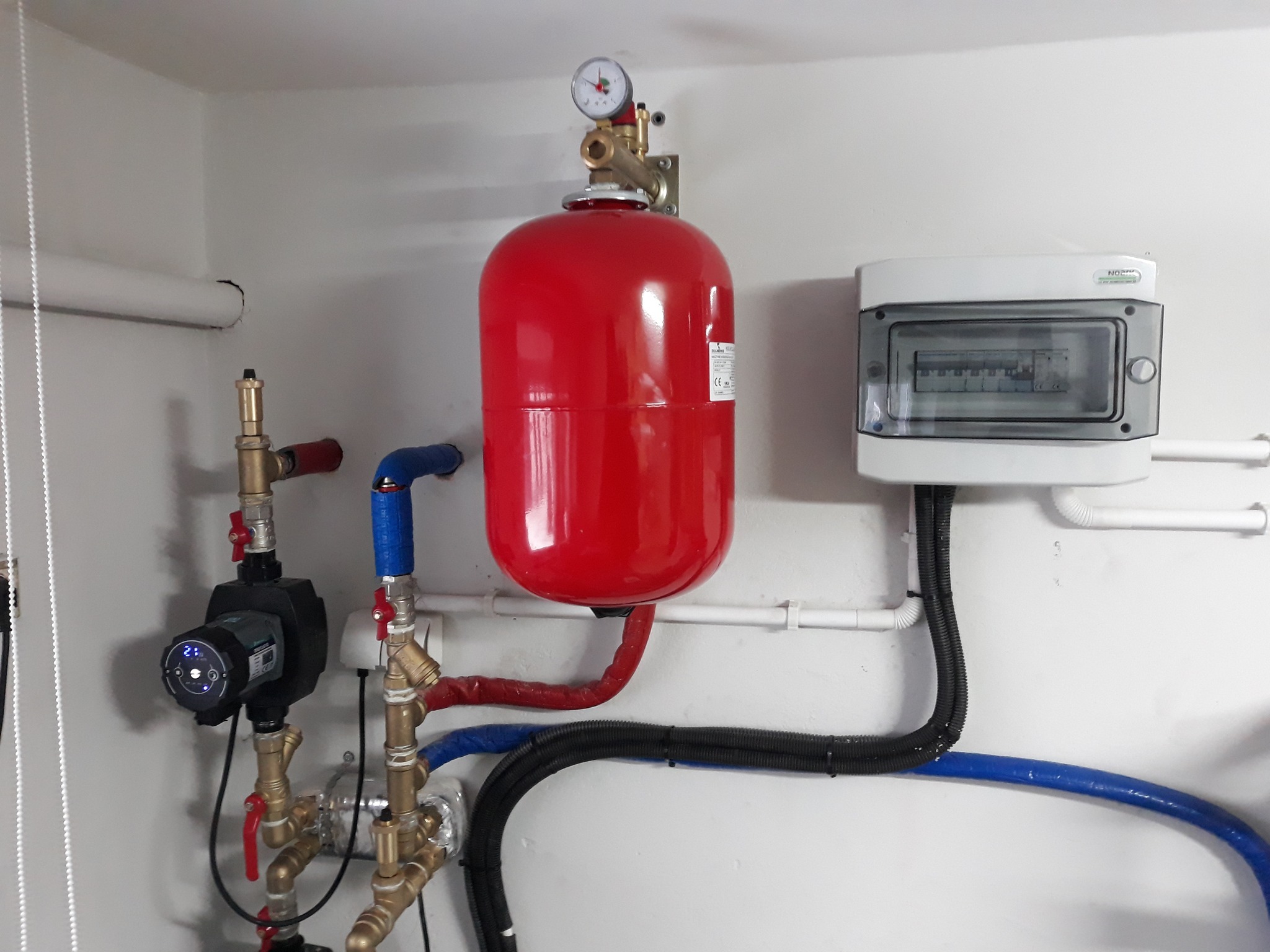Czerwony zbiornik ciśnieniowy z manometrem, pompa obiegowa z wyświetlaczem, rury miedziane i plastikowe w instalacji hydraulicznej, skrzynka elektryczna z bezpiecznikami na białej ścianie.