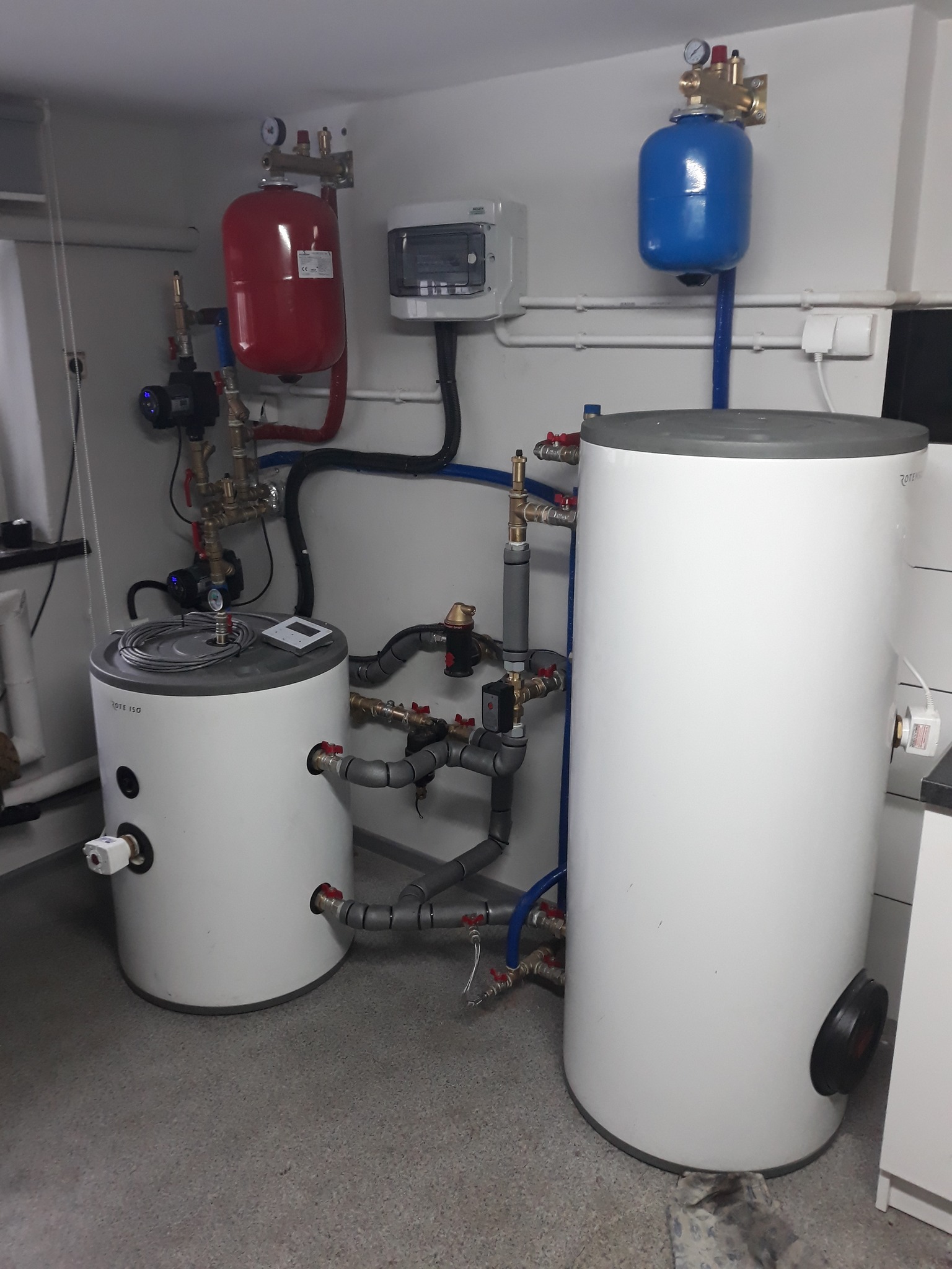 Dwa białe zbiorniki akumulacyjne Rotenso z podłączeniami hydraulicznymi, czerwony i niebieski zbiornik wyrównawczy ciśnienia zamontowane na ścianie, szara podłoga i białe ściany w kotłowni.