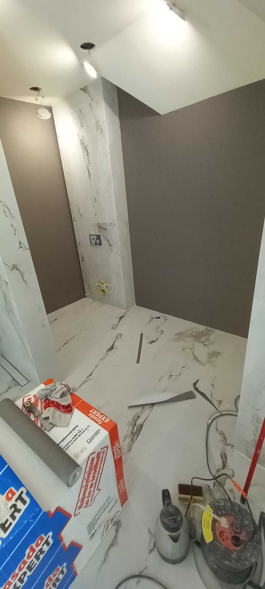 Wnętrze łazienki w trakcie wykańczania: marmurowe płytki na ścianach i podłodze, fragment ściany pomalowany na szaro, widoczne narzędzia i materiały budowlane, w tym paczka styropianu i rolka folii.
