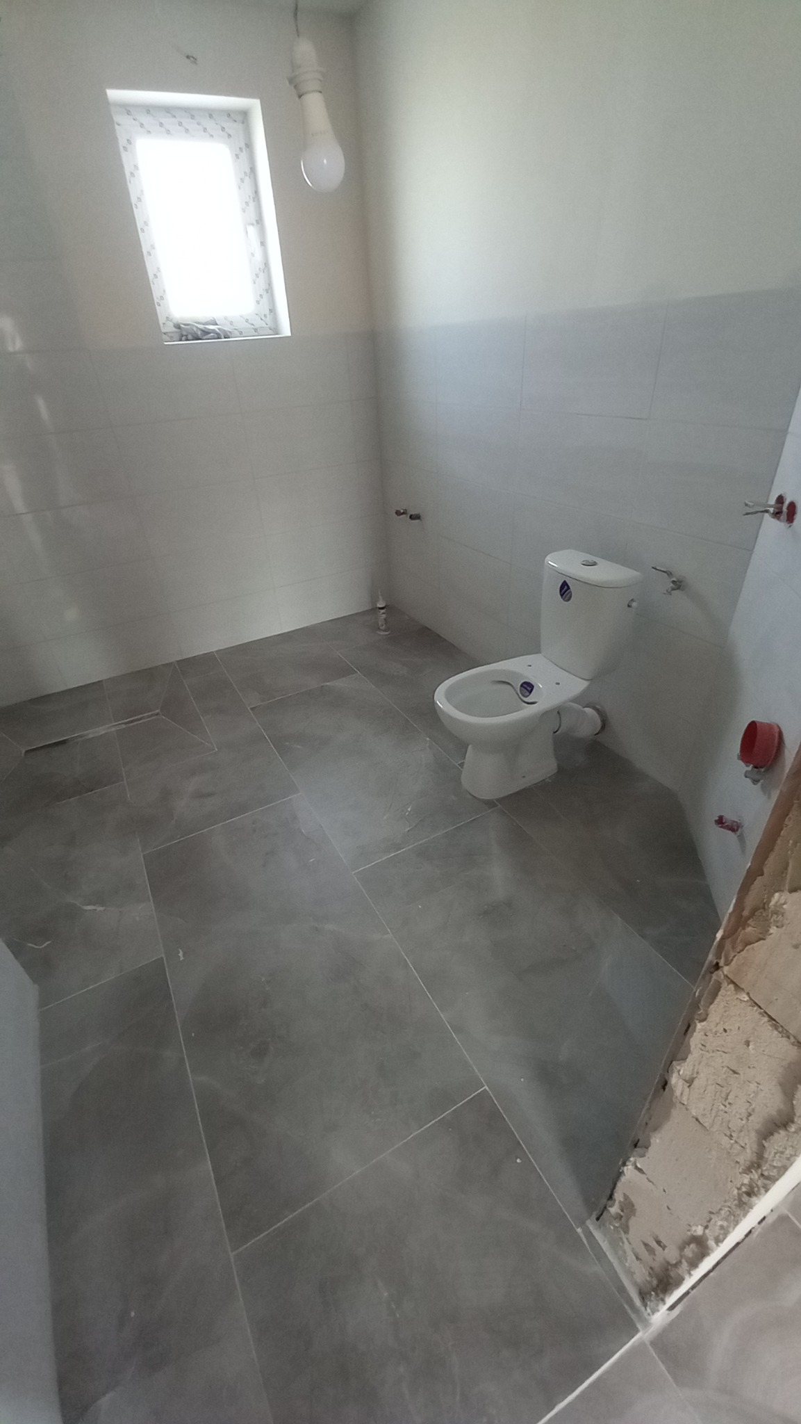 Nowa toaleta z szarymi płytkami podłogowymi i białymi płytkami ściennymi, widoczne niedokończone obrzeża przy drzwiach i surowa żarówka.