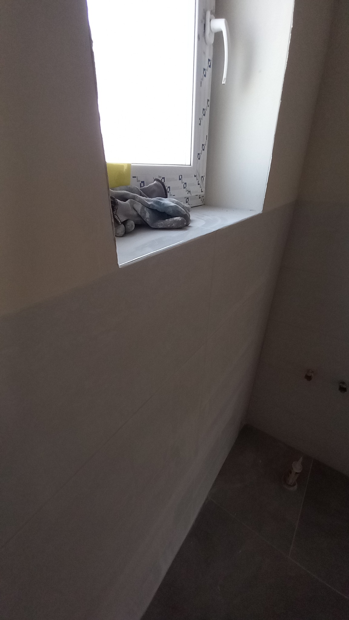 Świeżo wykafelkowana ściana w łazience z widocznym oknem, na parapecie leżą rękawice robocze i gąbka, na podłodze ciemne płytki.