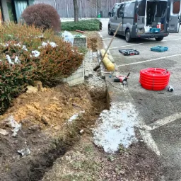 Wykopany rów w ziemi obok parkingu, widoczne narzędzia, zwój czerwonego kabla, samochód dostawczy z otwartymi drzwiami i skrzynkami narzędziowymi wewnątrz.