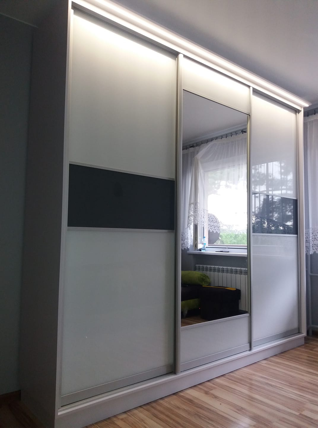 Biało-szara szafa przesuwna z lustrem, widoczna w pokoju z drewnianą podłogą i oknem z firanką.
