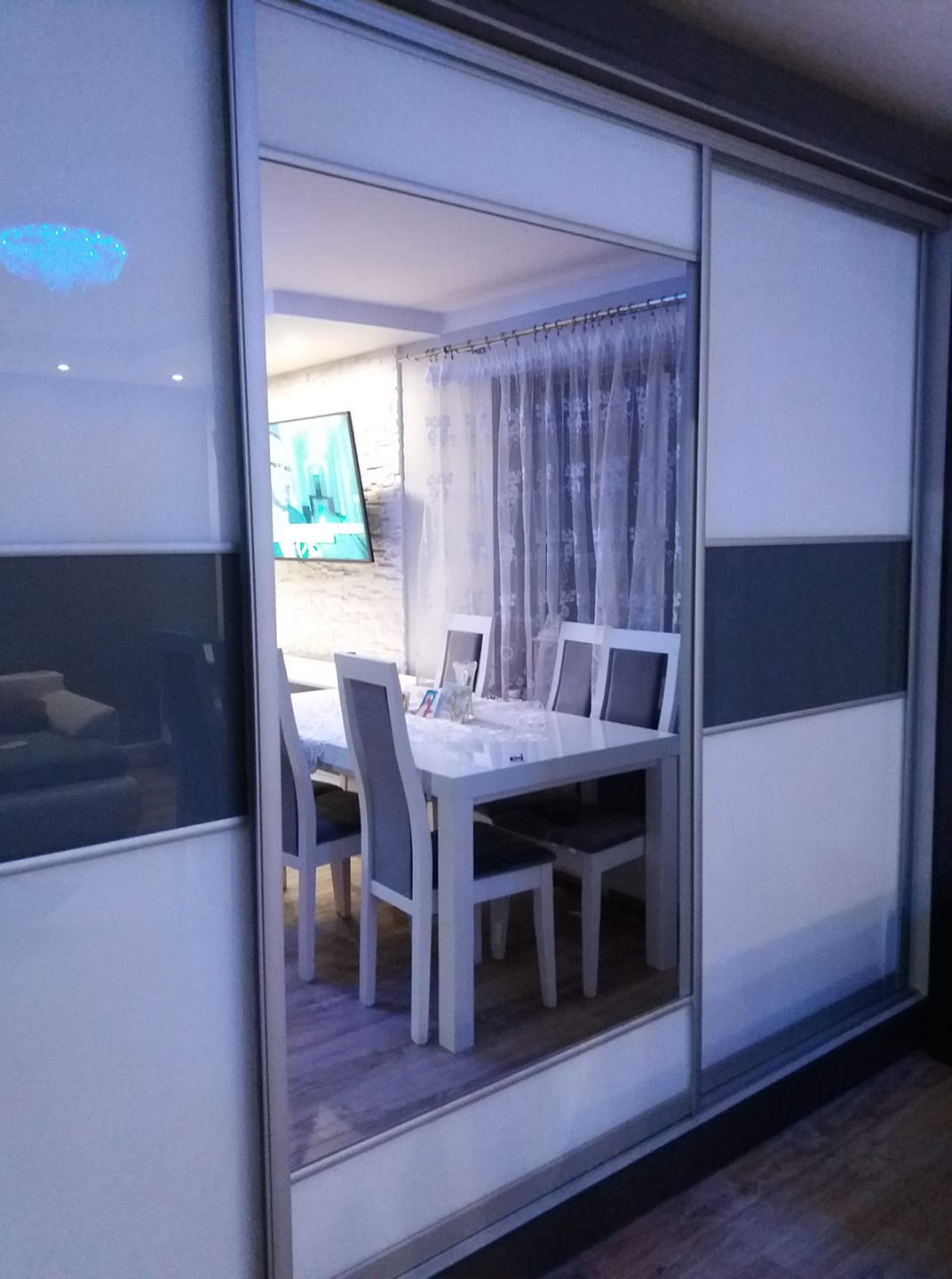 Nowoczesna szafa przesuwna z lustrzanym panelem, odbijająca wnętrze jadalni z białym stołem i krzesłami w Łabiszynie. Widoczny telewizor na ścianie z dekoracyjnej cegły oraz delikatne, białe zasłony.