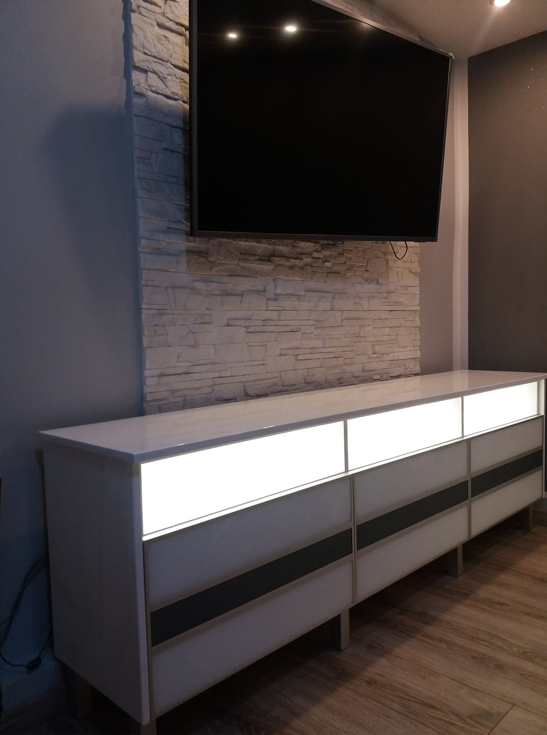 Nowoczesna szafka RTV z podświetlanymi frontami szuflad, umieszczona pod telewizorem na ścianie wyłożonej dekoracyjnym kamieniem.