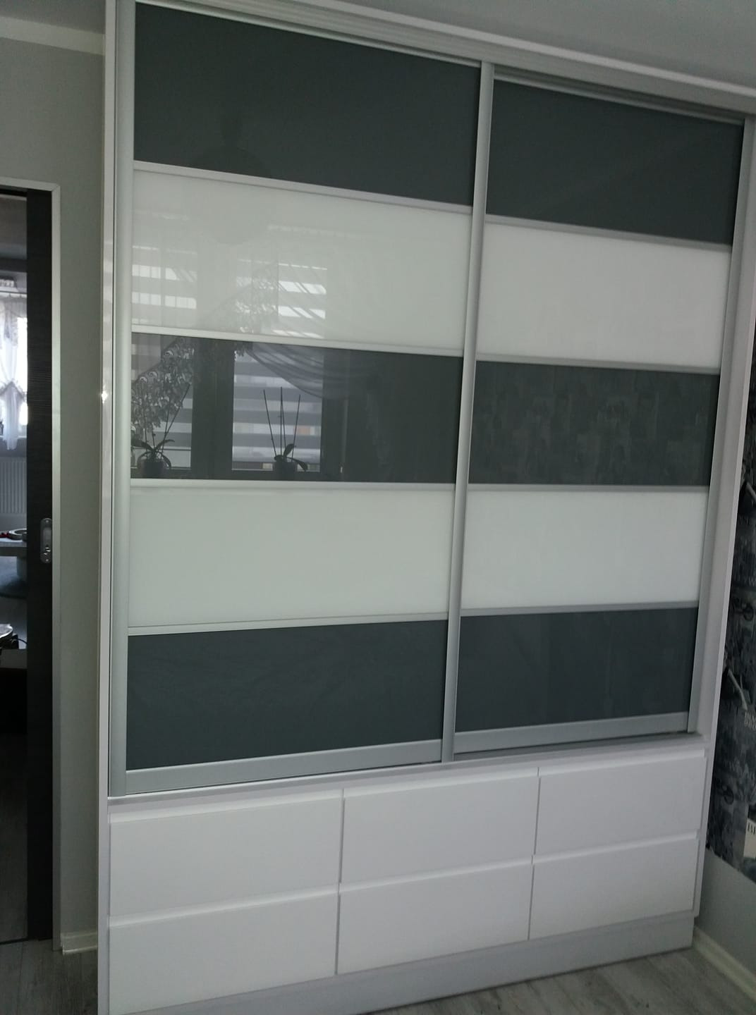 Biała szafa przesuwna z szarymi i białymi panelami, osadzona na komodzie z szufladami, widoczna z boku w pomieszczeniu.