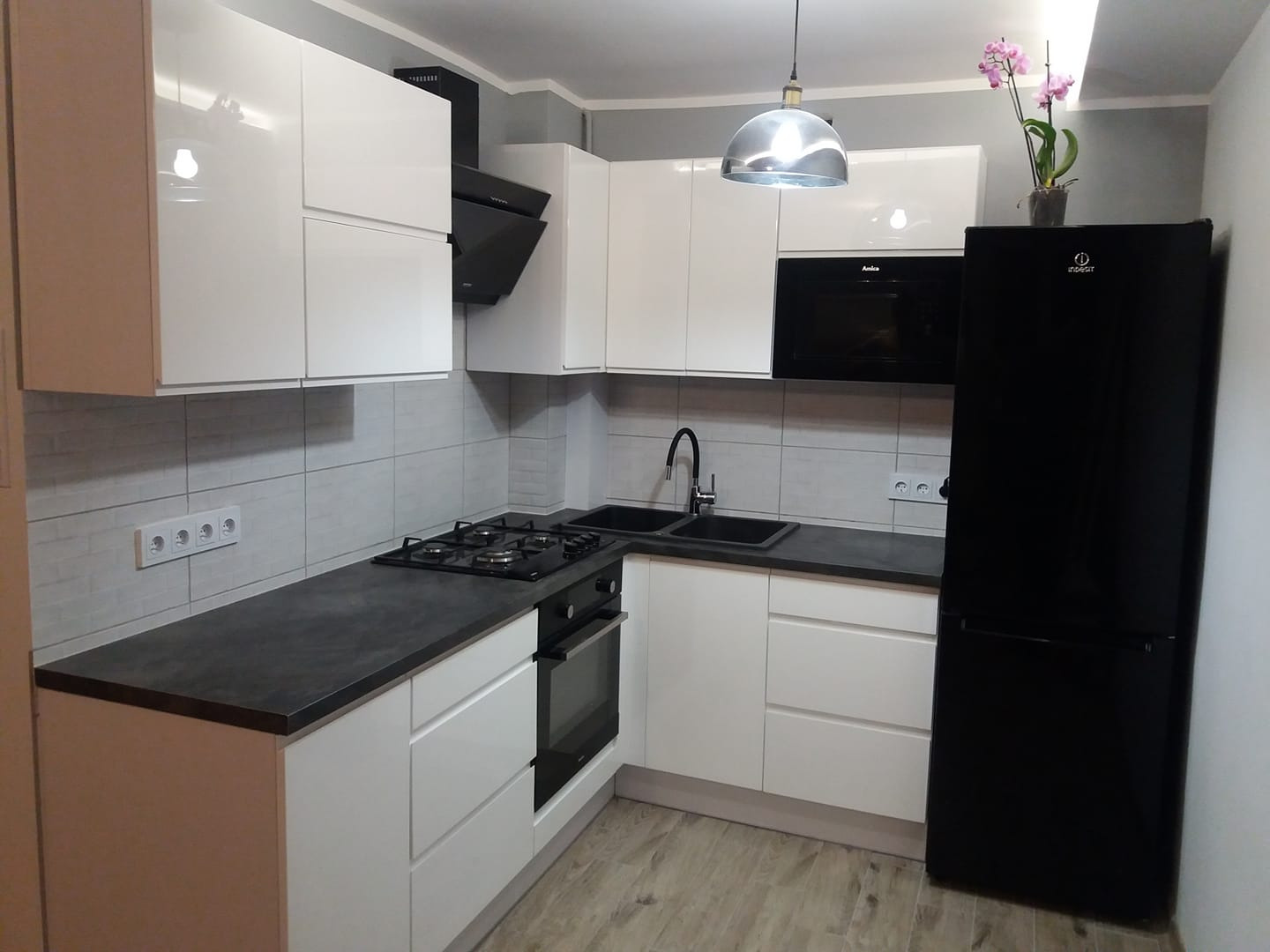 Nowoczesna kuchnia z białymi, lakierowanymi frontami, czarnym blatem i sprzętem AGD, oświetlona wiszącą lampą w stylu industrialnym i dopełniona storczykiem w doniczce.