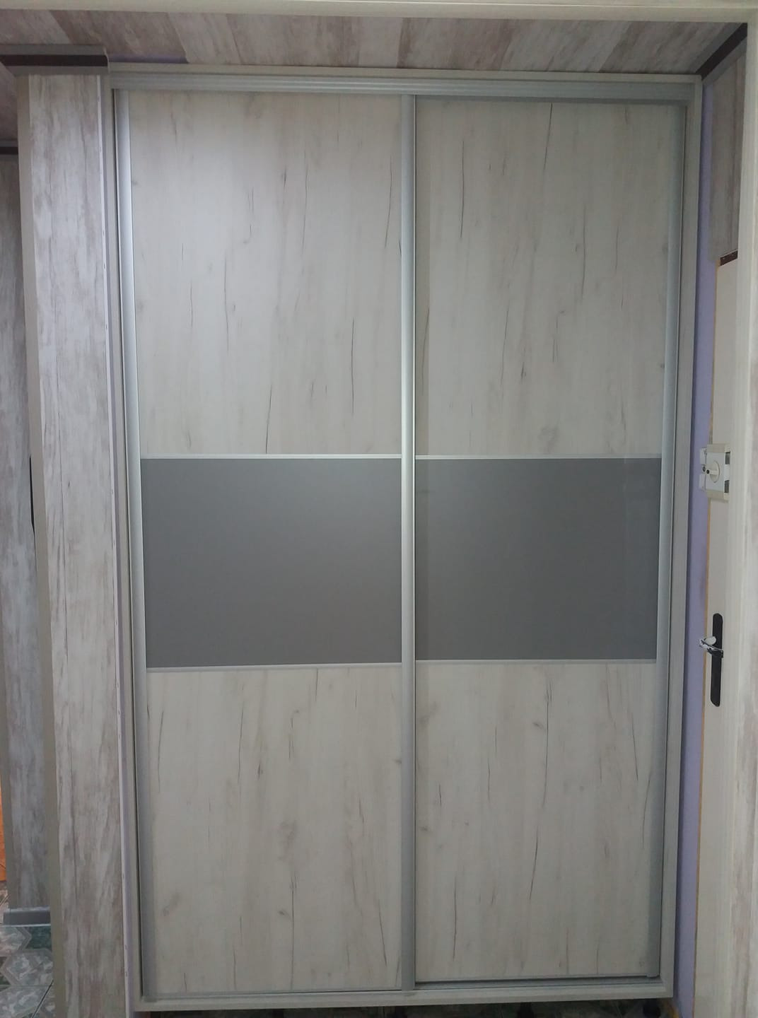 Dwudrzwiowa szafa przesuwna z panelami w kolorze jasnego drewna i szarymi pasami, widoczna w korytarzu z płytkami na podłodze.