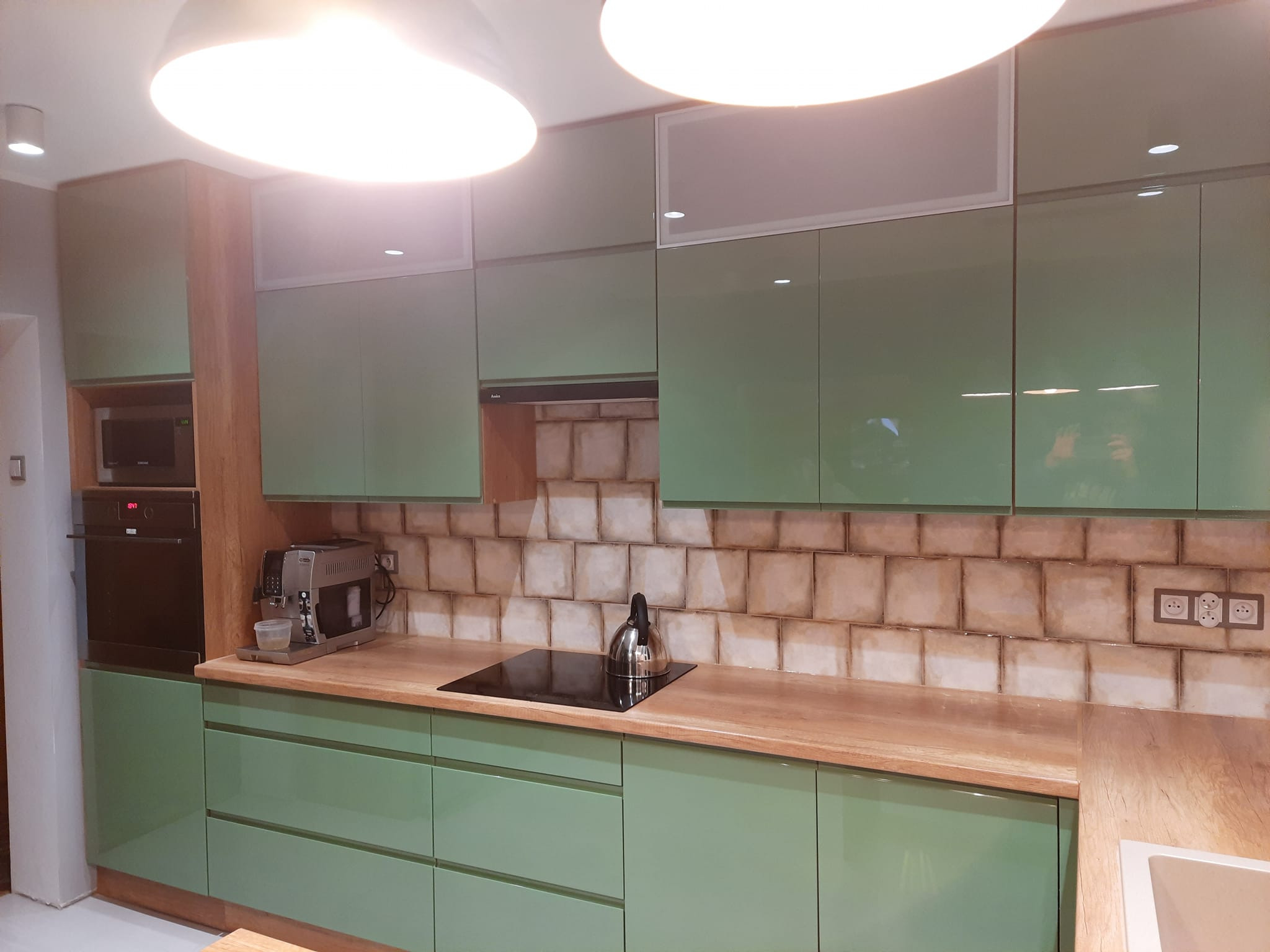 Nowoczesna kuchnia z zielonymi frontami szafek, drewnianym blatem i płytkami imitującymi cegłę na ścianie, wyposażona w sprzęt AGD.