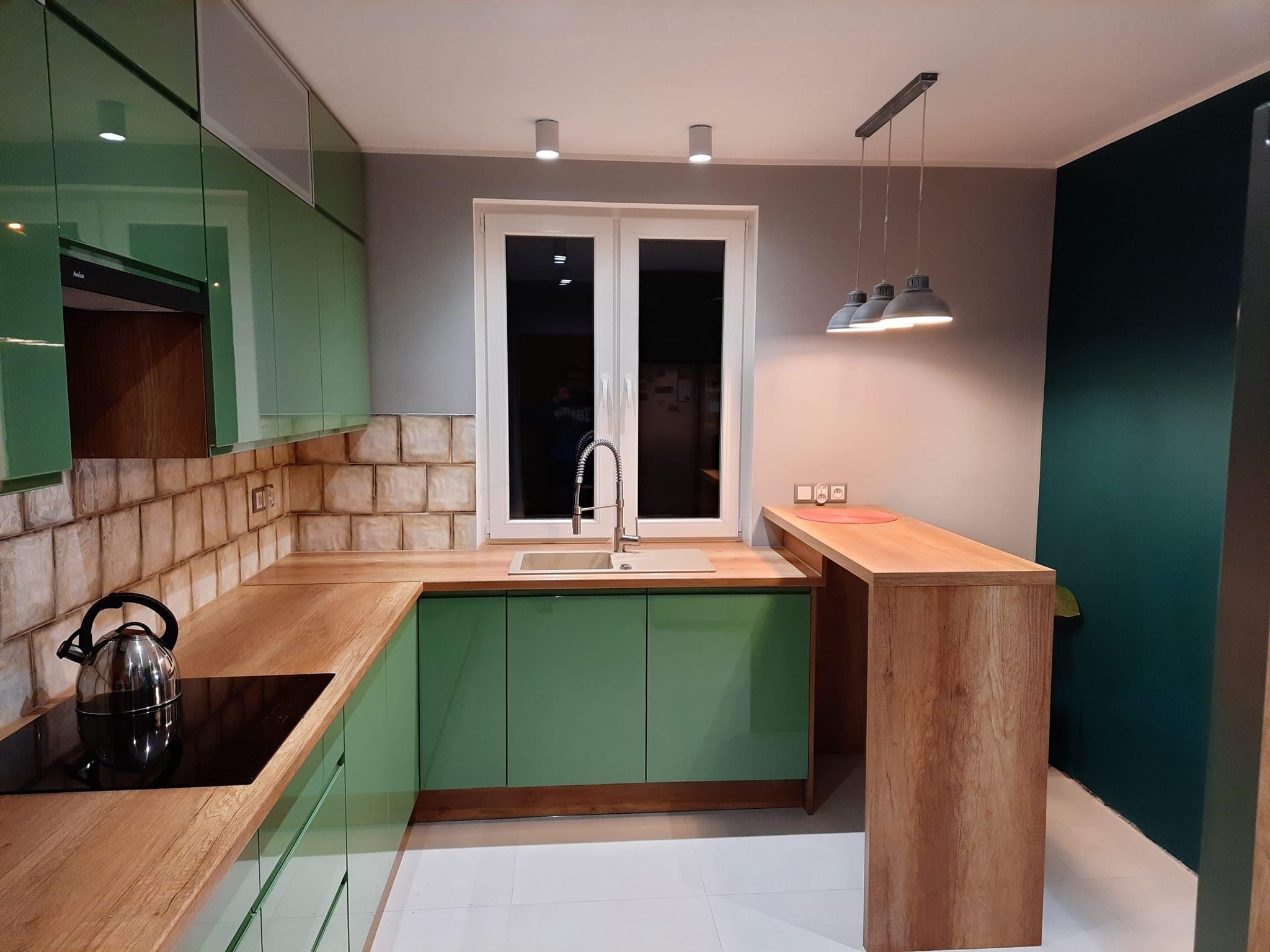 Nowoczesna kuchnia z zielonymi frontami, drewnianym blatem i barkiem, oświetlona trzema lampami wiszącymi, z czajnikiem na płycie indukcyjnej.