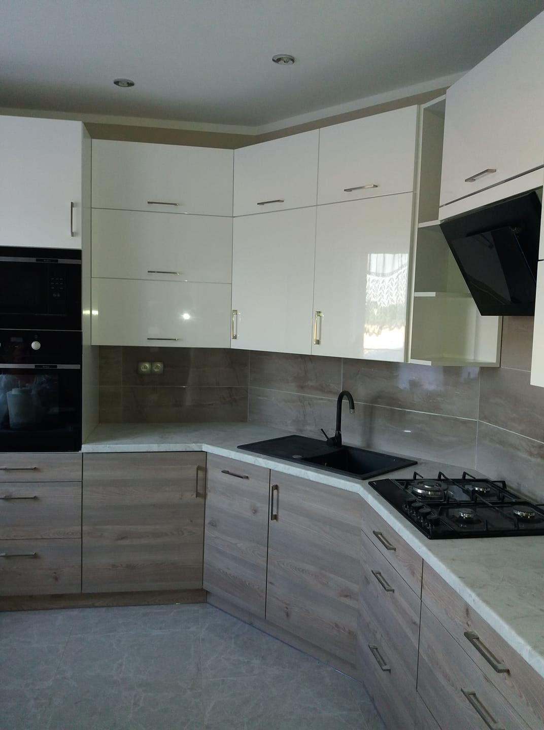 Nowoczesna kuchnia z białymi, lakierowanymi szafkami górnymi i dolnymi w odcieniach drewna, czarny zlew i płyta gazowa, szare płytki na ścianie i podłodze.