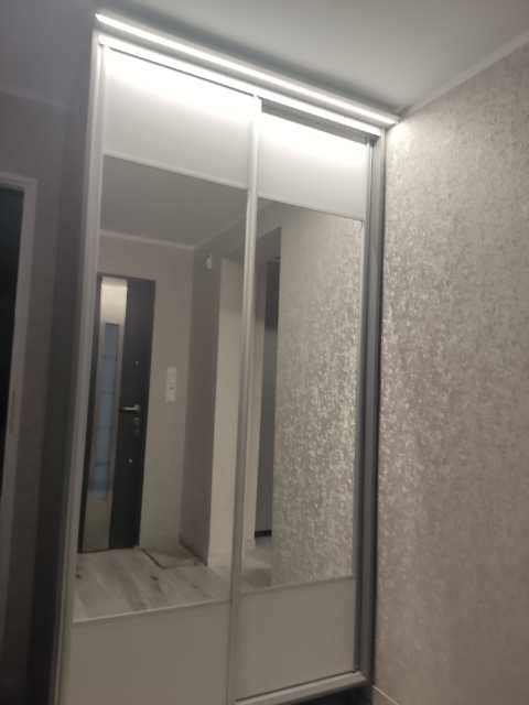 Wbudowana szafa z przesuwnymi drzwiami z lustrami i białymi panelami, widok na wnętrze mieszkania przez otwarte drzwi.